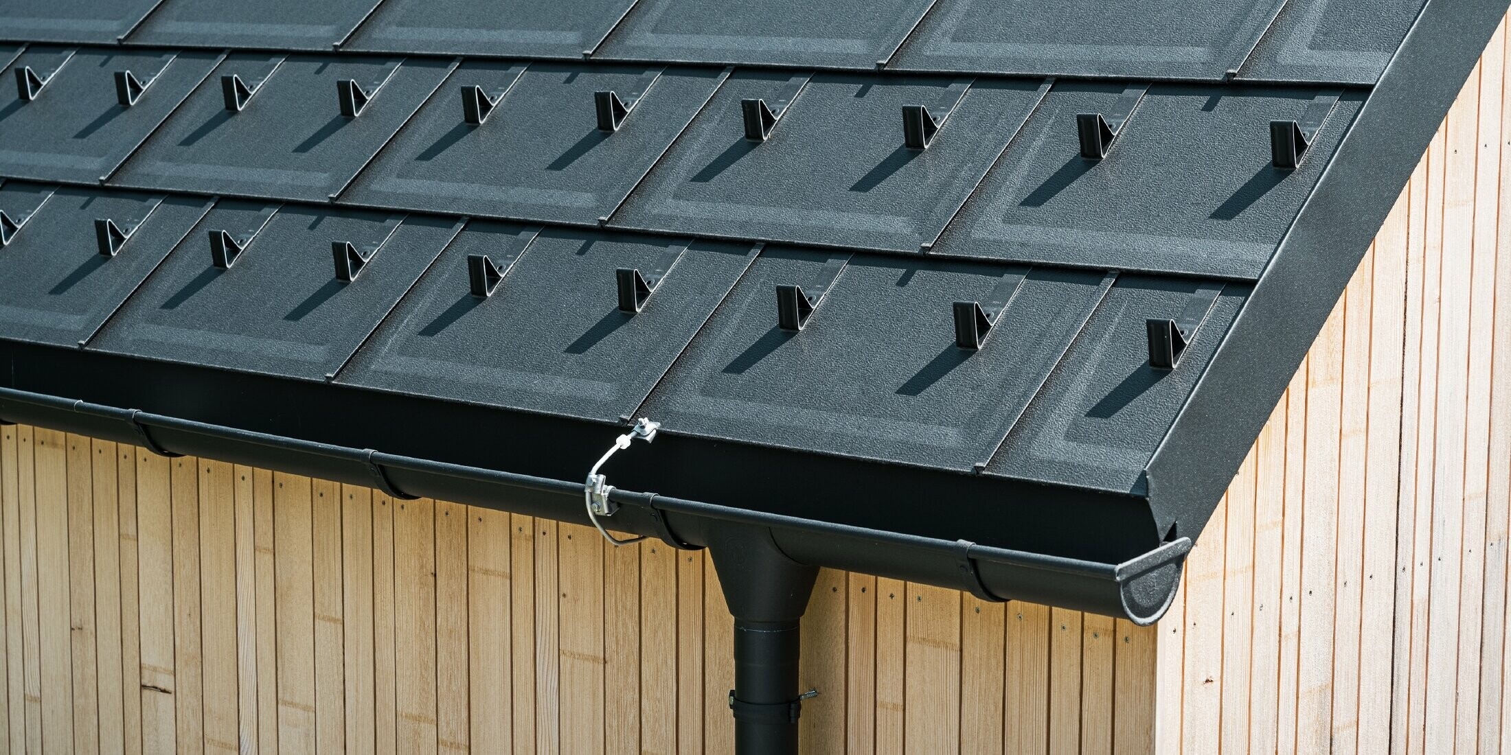 Primo piano della tegola R.16 PREFA con nasi fermaneve, canale di gronda e tubo pluviale nel colore nero P.10 con parafulmine e facciata in legno verticale