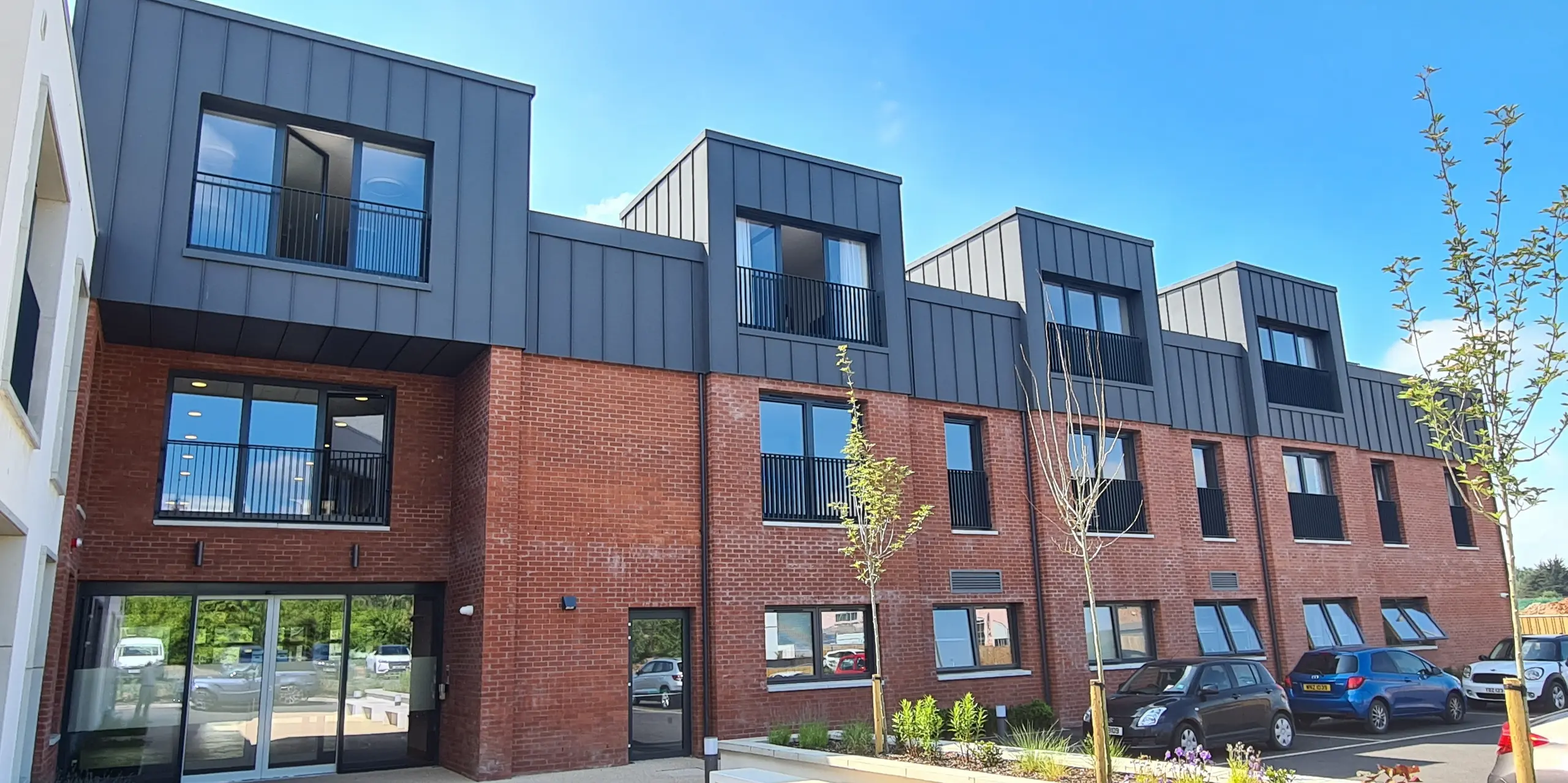 Vue de face de Harberton Hall à Belfast, en Irlande du Nord. La toiture à joint debout en PREFALZ P.10 gris sombre complète parfaitement la façade en briques. De grandes fenêtres et des lucarnes à toit plat soulignent la conception innovante du bâtiment de vie assistée et créent une atmosphère accueillante. La porte d'entrée vitrée donne un aperçu de l'intérieur de l'établissement.