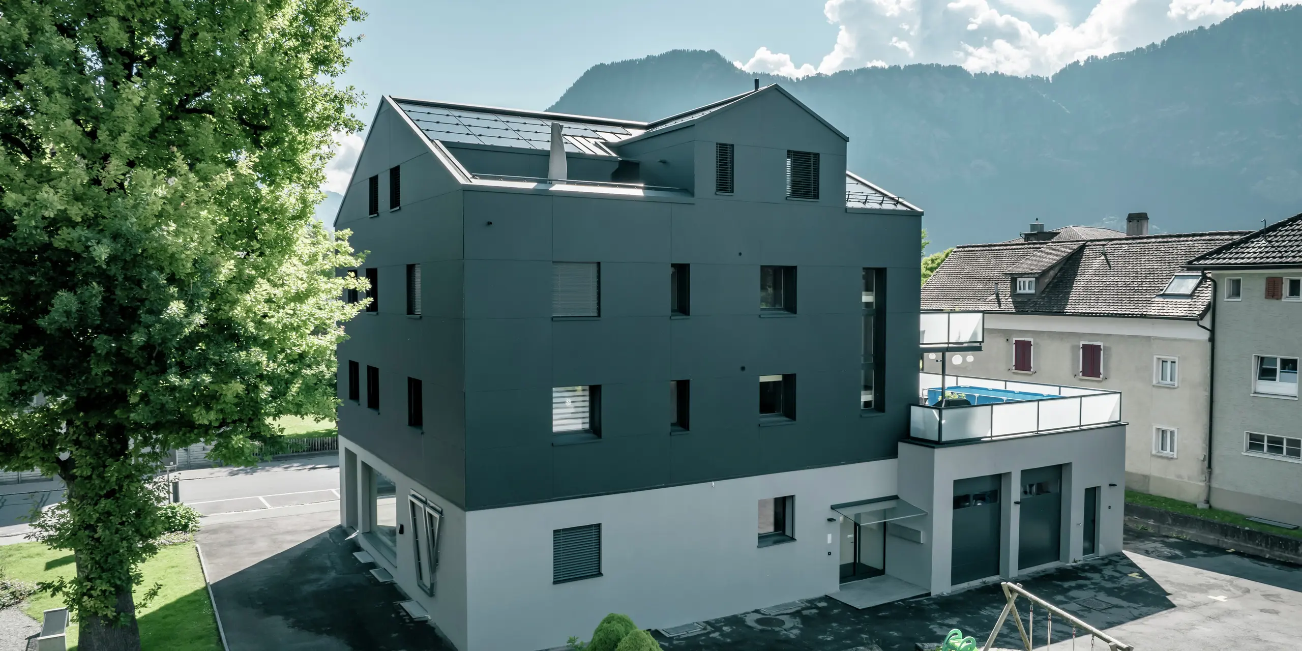 Bâtiment résidentiel et commercial modernisé avec une façade PREFA de couleur anthracite en panneaux composites en aluminium PREFABOND. La vue latérale montre des lignes épurées et un design contemporain. Au premier plan se trouvent des espaces verts bien entretenus et une petite aire de jeux. En arrière-plan, vous pouvez voir des montagnes et un ciel ensoleillé avec des nuages ​​légers.