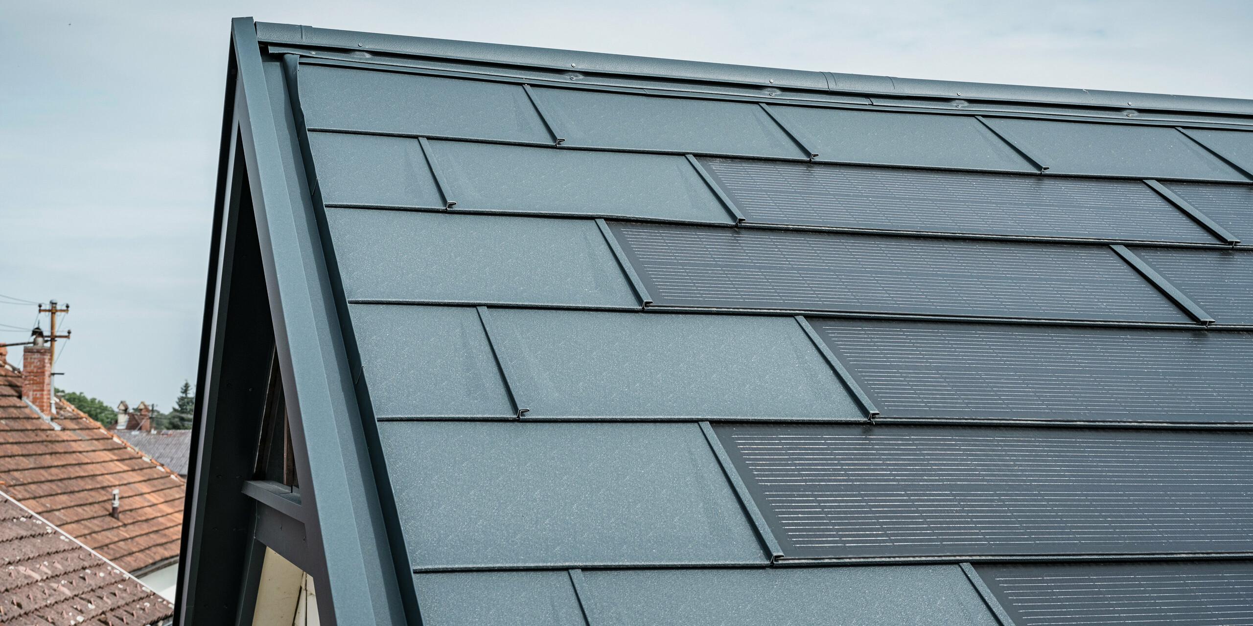 Vista dettagliata del tetto di una casa dotata dell'innovativa tegola PREFA Solar. Le tegole con celle fotovoltaiche integrate si presentano in un elegante colore antracite. La superficie omogenea si integra perfettamente nel tetto, creando un aspetto moderno e pulito. L'innovativo sistema di copertura garantisce un utilizzo efficiente dell'energia senza compromettere l'estetica.