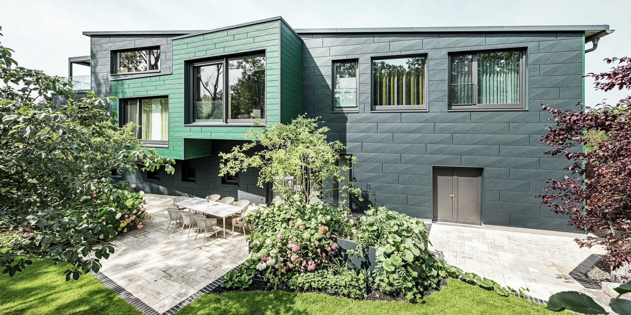La casa unifamiliare a Türkheim colpisce per la sua facciata straordinaria, rivestita con pannelli PREFA Siding X nei colori antracite e verde muschio. Le linee pulite e la scelta cromatica decisa dei pannelli in alluminio conferiscono all'edificio un aspetto moderno ed elegante. In primo piano si vede l'ampio giardino con un'elegante terrazza e un tavolo da pranzo con sedie moderne. Aiuole colorate e piante rigogliose creano un vivace contrasto verde con la robusta facciata. La facciata in alluminio, resistente e di facile manutenzione, protegge l'edificio e garantisce una bellezza senza tempo.