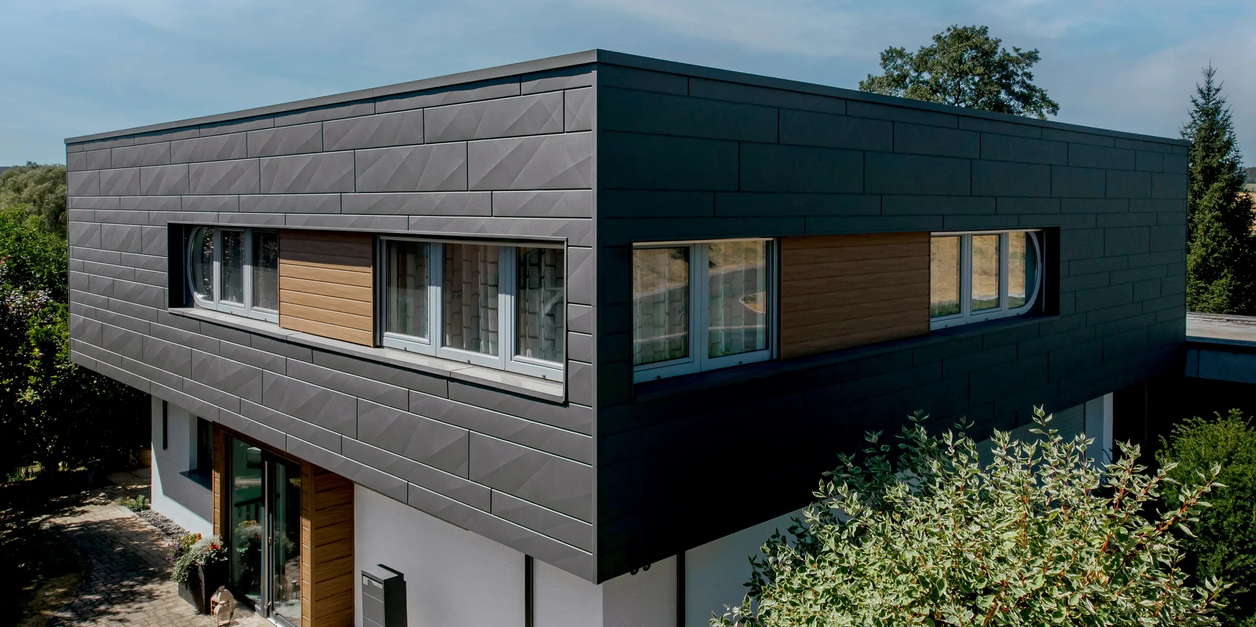 Modernes Einfamilienhaus in Marksuhl mit markanter PREFA Aluminiumfassade: Die großflächigen PREFA Siding.X Paneele in P.10 Dunkelgrau verleihen dem auskragenden Baukörper eine klare, geometrische Optik. Akzentflächen mit PREFA Siding in Walnuss braun schaffen einen warmen Kontrast. Die langlebige Aluminiumverkleidung bietet zuverlässigen Wetterschutz und eine dauerhaft elegante Gebäudehülle.