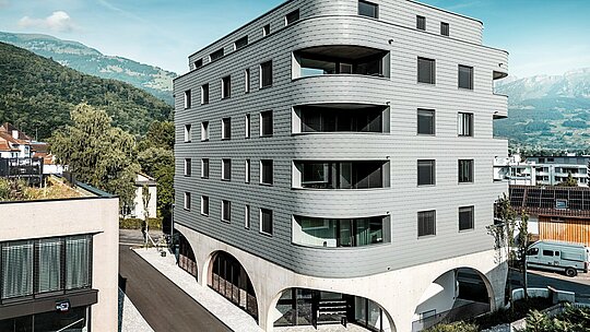 Außenansicht des Bernerhauses in Buchs mit markanter, dreieckiger Form und weich gerundeten Balkonbändern. Die Fassade aus PREFA Wandschindeln in P.10 Hellgrau umhüllt das Gebäude wie eine schuppenartige Aluminiumhaut und erzeugt ein präzises, lebendiges Fassadenbild. Über dem massiven Betonsockel mit eleganten Arkaden wirkt die leichte, witterungsbeständige PREFA Aluminiumfassade besonders ausdrucksstark. Langlebig, wartungsarm und formstabil setzt sie vor alpiner Bergkulisse ein modernes architektonisches Statement.