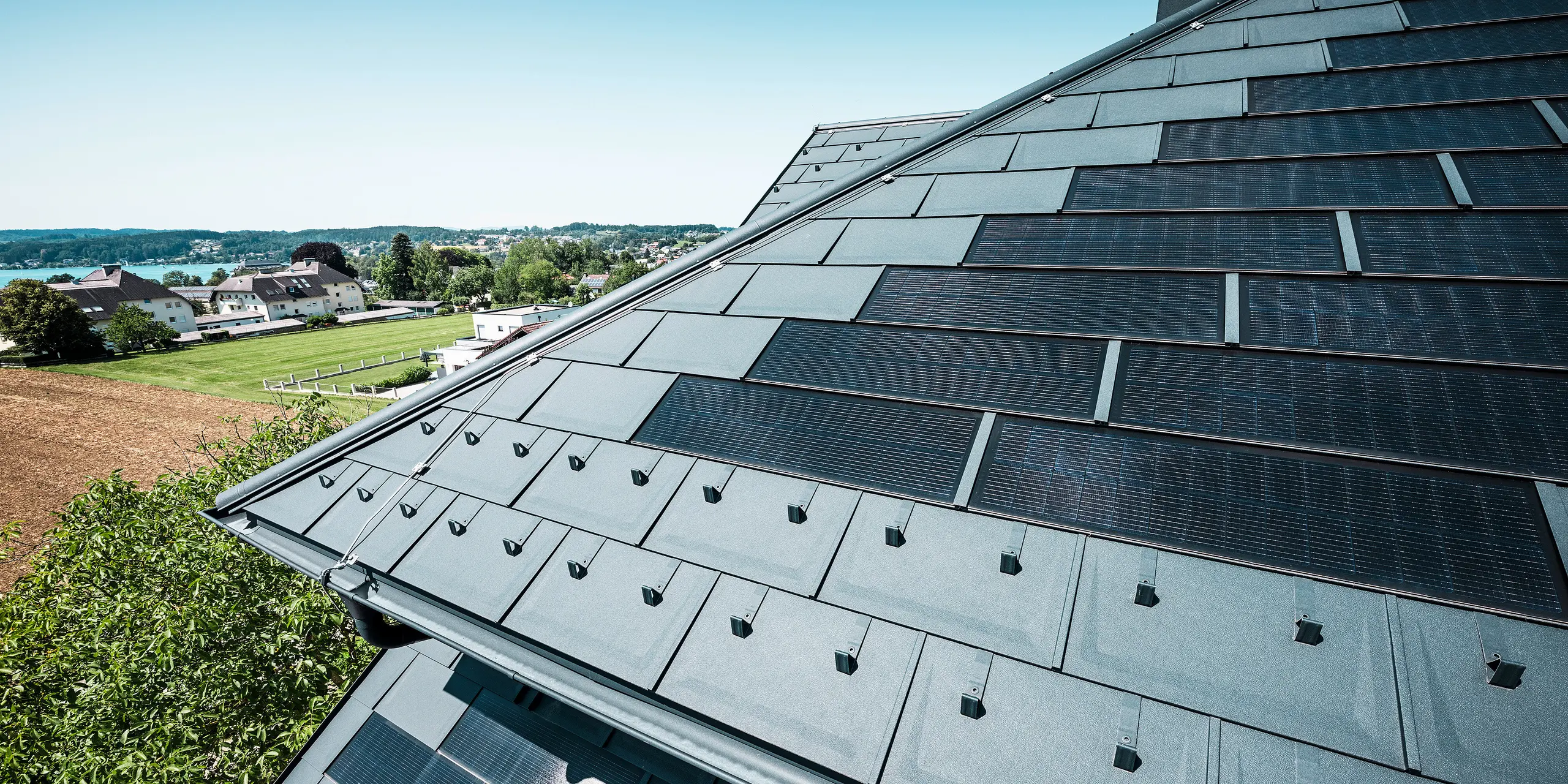 Nahaufnahme des neu sanierten Daches der Zenz'n Stub'n in Schörfling am Attersee, eingedeckt mit langlebigen PREFA Dachplatten R.16 und innovativen PREFA Solardachplatten in der Farbe P.10 Anthrazit. Das Aluminiumdach verbindet modernes Design mit nachhaltiger Energiegewinnung. Die robuste Dachentwässerung in passendem P.10 Anthrazit fügt sich harmonisch in das Gesamtbild ein. In der Ferne ist im Hintergrund der blaue Attersee zu erkennen.