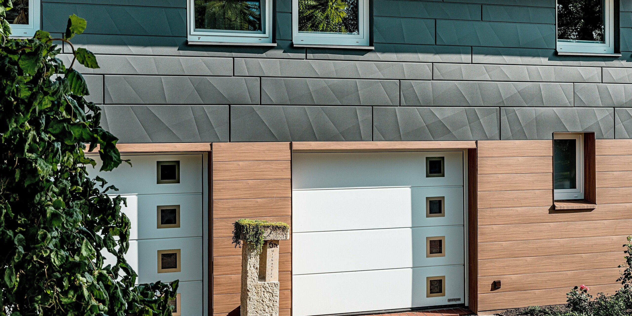 Detailansicht der sanierten Fassade eines Einfamilienhauses mit hochwertiger PREFA Aluminiumverkleidung. Im Fokus stehen die weißen Garagentore, eingerahmt von PREFA Siding in Walnussbraun mit authentischer Holzoptik, die dem Eingangsbereich Wärme und Tiefe verleiht. Darüber setzt PREFA Siding.X in P.10 Dunkelgrau mit markanten Längs- und Querkantungen einen modernen Akzent und erzeugt ein lebendiges Spiel aus Licht und Schatten. Die klare horizontale Linienführung, kombiniert mit langlebigen, witterungsbeständigen und pflegeleichten Aluminiumpaneelen, macht diese Fassadensanierung zu einem eindrucksvollen Beispiel für zeitgemäße Architektur und nachhaltige Qualität.