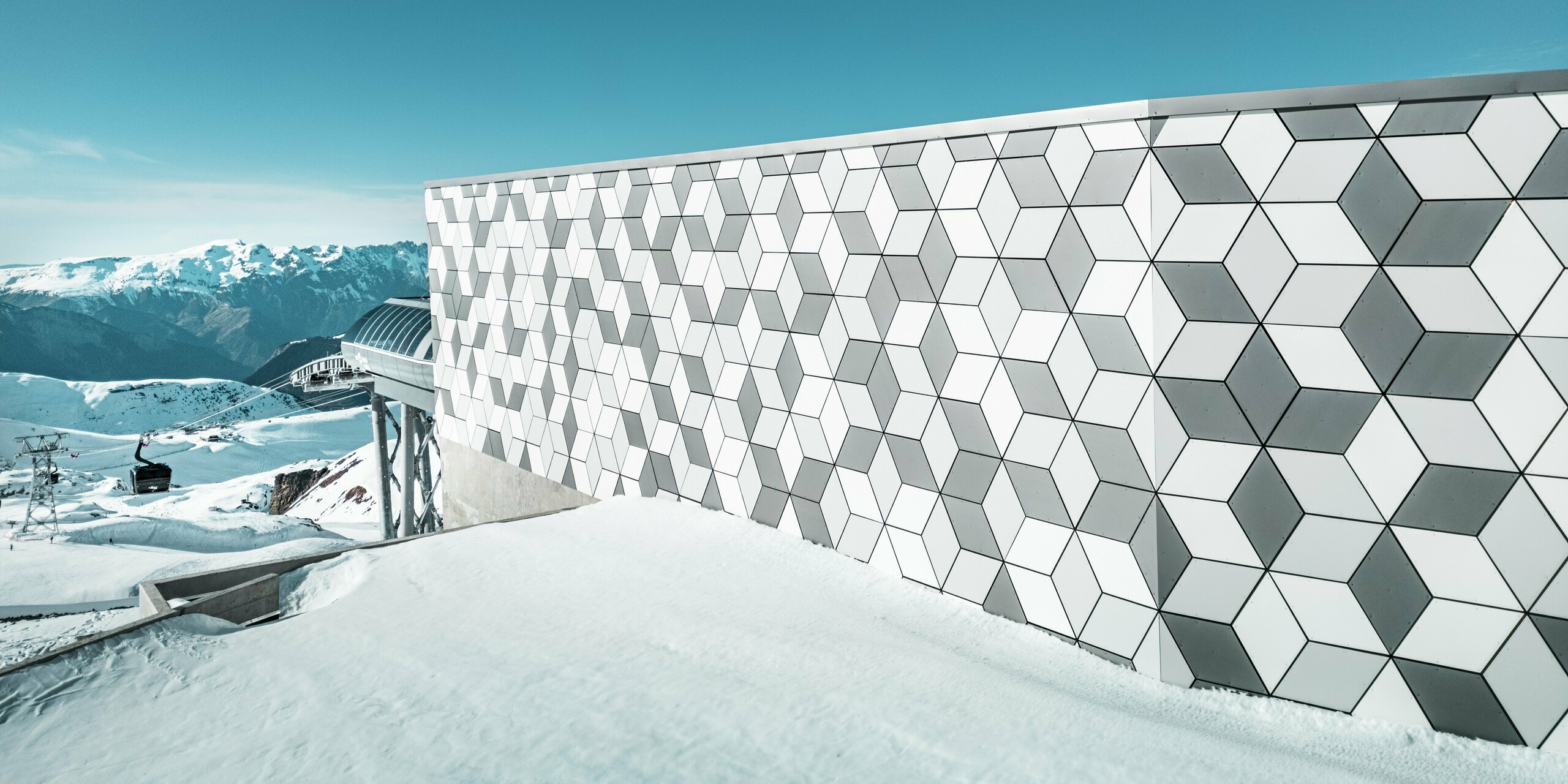 Aufnahme der Aluminiumfassade der Seilbahnstation in Les Deux Alpes: Die markante Fassade aus PREFABOND Aluminiumverbundplatten beeindruckt mit ihrer auffälligen, geometrischen Rautenmusteranordnung. Die harmonische Kombination aus Weiß und Grau erzeugt einen modernen Look, der sich nahtlos in die majestätische Berglandschaft einfügt. Hinter der Fassade sind der strahlend blaue Himmel und das majestätische Bergpanorama zu sehen.