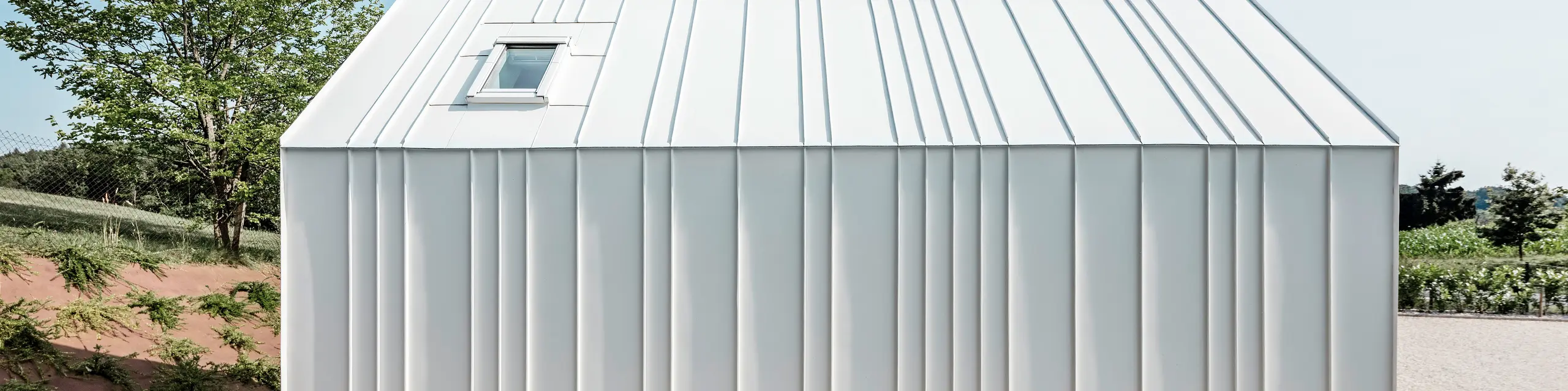 Seitenansicht von einem der Tiny Häuser 'Pri Momi' in Kančevci, Slowenien. Das Mikrohaus ist umhüllt von einer eleganten Stehfalzdeckung aus dem langlebigen Dach- und Fassadensystem PREFALZ. Die hochwertige Blechverkleidung ist in der Farbe P.10 Reinweiß ausgeführt. Das Bild zeigt die präzise ausgeführten Falze und die eleganten Linien der weißen Aluminiumhülle, die dem Gebäude ein modernes und minimalistisches Aussehen verleihen. Die natürliche Umgebung und das klare, schlichte Design harmonieren perfekt miteinander.