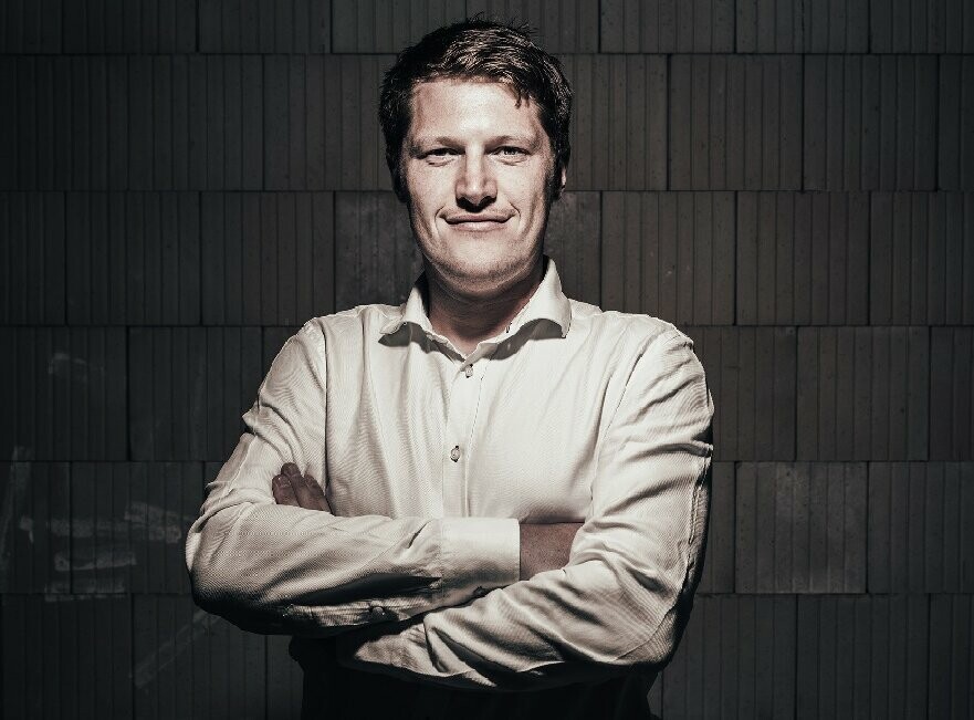 Portrait de l’architecte Andreas Gruber. Il porte une chemise blanche et croise les bras face à l’appareil photo, ce qui lui donne un air déterminé.