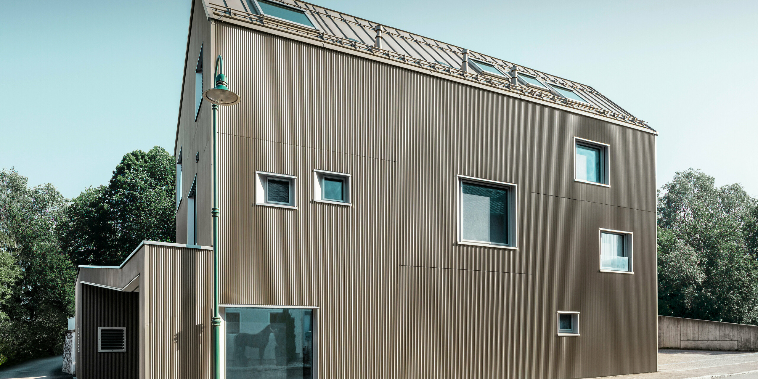Blick von der Straße auf ein zweistöckiges Einfamilienhaus in Frankenburg, Österreich, gekennzeichnet durch ein PREFA Komplettsystem. Das Dach wurde mit PREFALZ in Bronze eingedeckt.. Die Fassade präsentiert sich in einer subtilen Textur, die durch die vertikalen Linien des Zackenprofils entsteht und ein harmonisches Schattenspiel erzeugt. Das Haus besitzt eine asymmetrische Fensteranordnung, die den modernen und unkonventionellen Charakter des Designs betont. Der Anbau auf der linken Seite ist stilistisch an das Hauptgebäude angelehnt, wodurch ein stimmiges Gesamtbild entsteht. Die klaren, sauberen Linien der Architektur kontrastieren wirkungsvoll mit dem blauen Himmel und den Bäumen im Hintergrund.