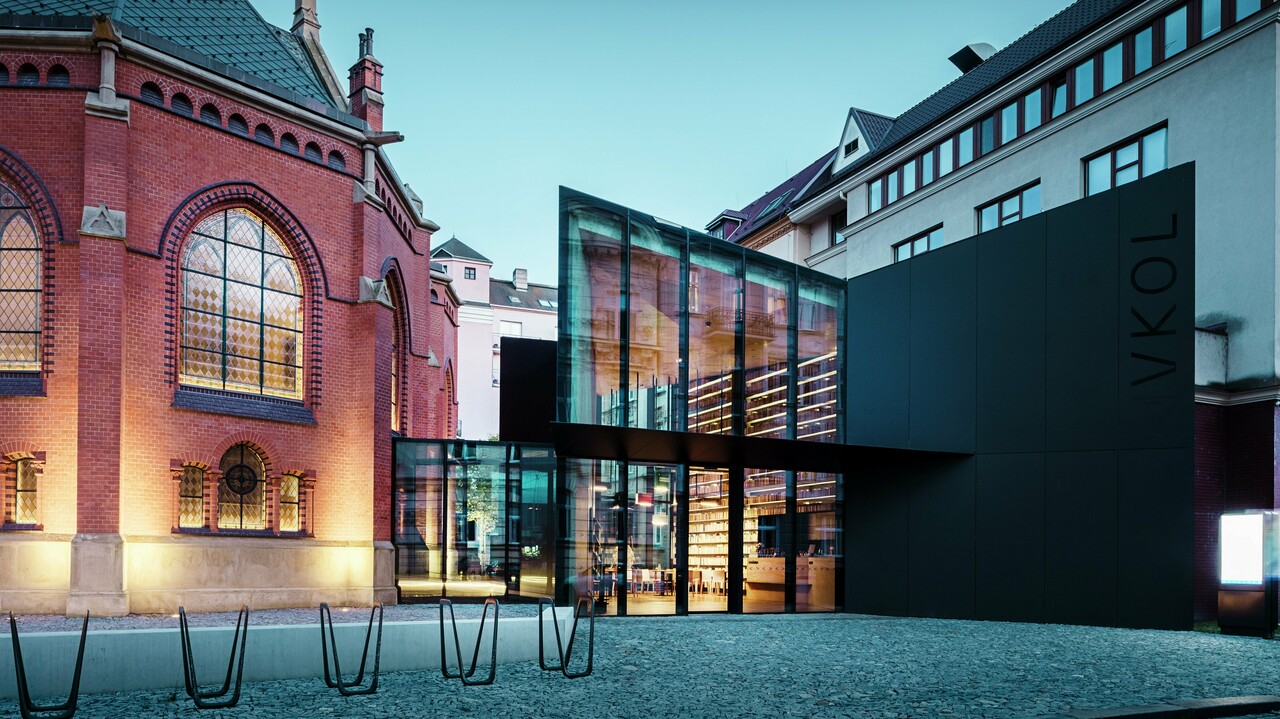 Das Bild zeigt eine beeindruckende Architektur in Olmütz, die die historische Rote Kirche mit einem modernen Neubau verbindet. Die prägnante schwarze Fassade aus PREFABOND Aluminium-Verbundplatten des Neubaus kontrastiert perfekt mit den roten Backsteinmauern der Kirche. Große Glasflächen ermöglichen einen Einblick in das Innere des Neubaus, wo Regale mit Büchern und eine einladende Atmosphäre zu sehen sind. Die präzise Ausführung der Materialien schafft eine harmonische Balance zwischen den historischen und modernen Elementen. Die Verwendung von langlebigen und ästhetisch ansprechenden Materialien wie PREFABOND sorgt nicht nur für eine moderne Optik, sondern auch für dauerhafte Qualität und Funktionalität. Diese Architektur zeigt auf eindrucksvolle Weise, wie Alt und Neu miteinander verschmelzen können, um ein einzigartiges urbanes Ensemble zu schaffen.