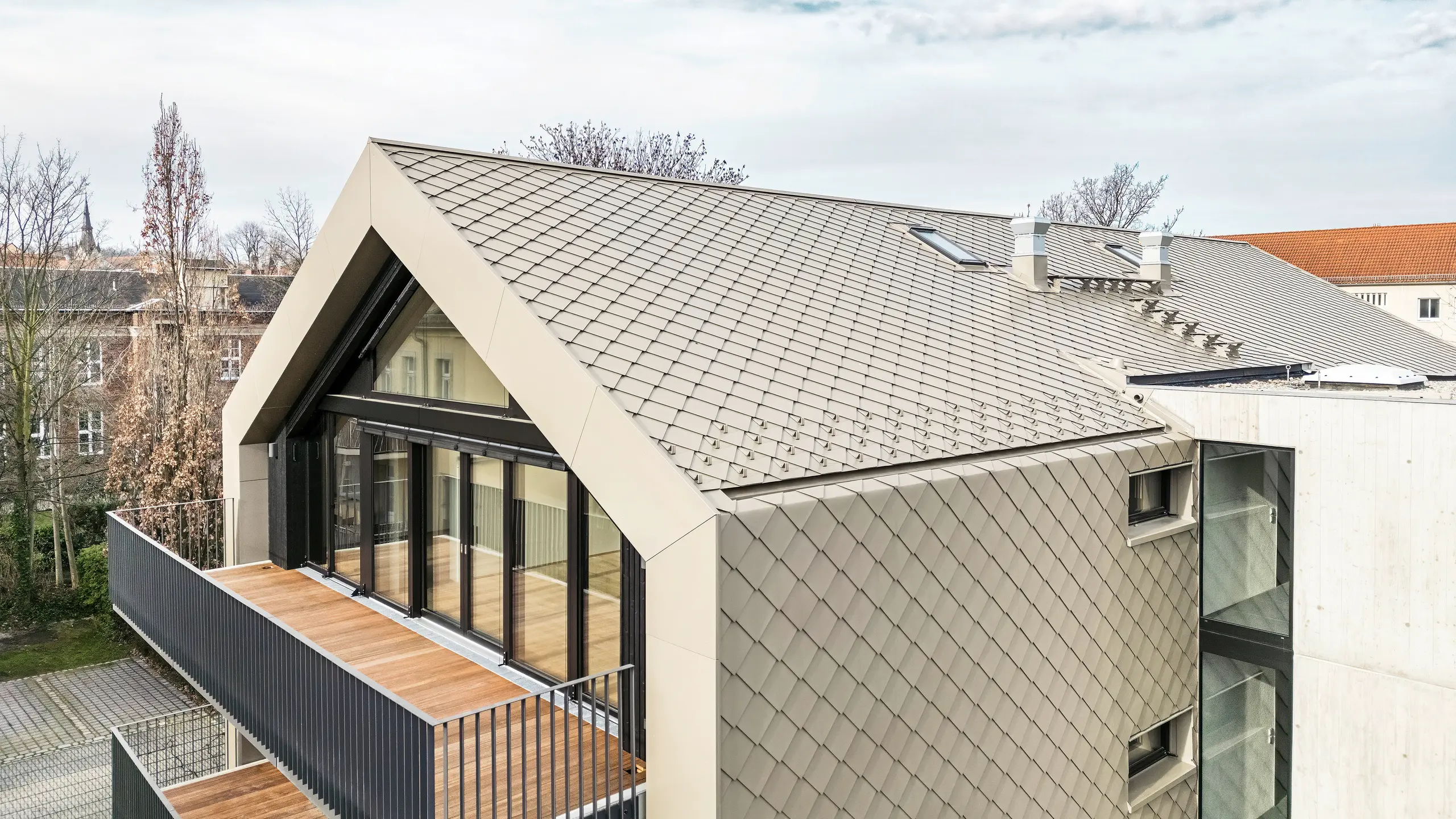 Vue plongeante d'un immeuble d'appartements moderne de la Chemnitzer Straße (Dresde) avec une enveloppe de bâtiment en losanges en aluminium PREFA 29 × 29 en bronze. L'image montre l'élégante charpente du toit, les grandes façades vitrées et les balcons ainsi que l'intégration harmonieuse du bâtiment dans l'environnement. Les matériaux de haute qualité soulignent l'architecture durable du bâtiment.