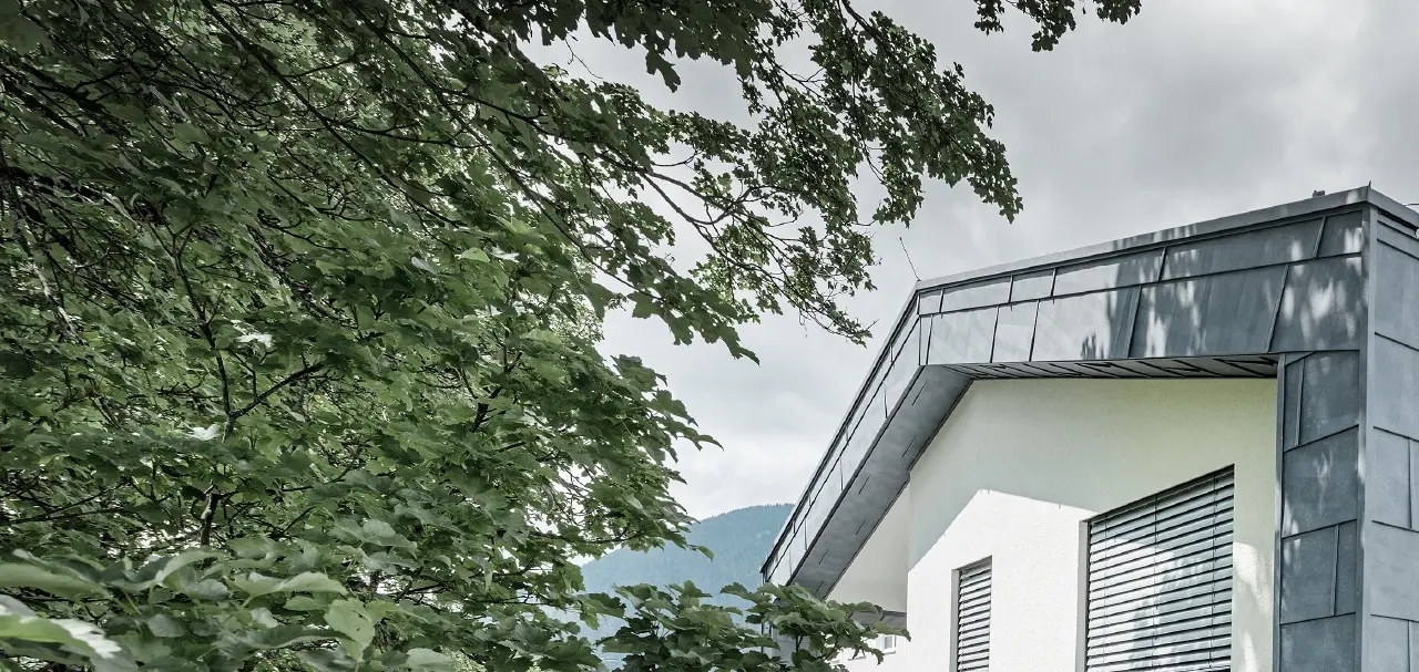 Moderna sopraelevazione di una casa con tetto piatto a due falde e senza sporto; il rivestimento della facciata è stato realizzato con il pannello per facciate FX.12 PREFA in grigio pietra.