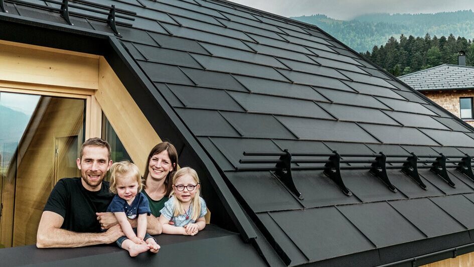 Die beiden Bauherren mit ihren Kindern genießen die Aussicht von der Loggia ihres neu erbauten Hauses, das mit stilvollen PREFA Solardachplatten in P.10 Schwarz ausgestattet ist.