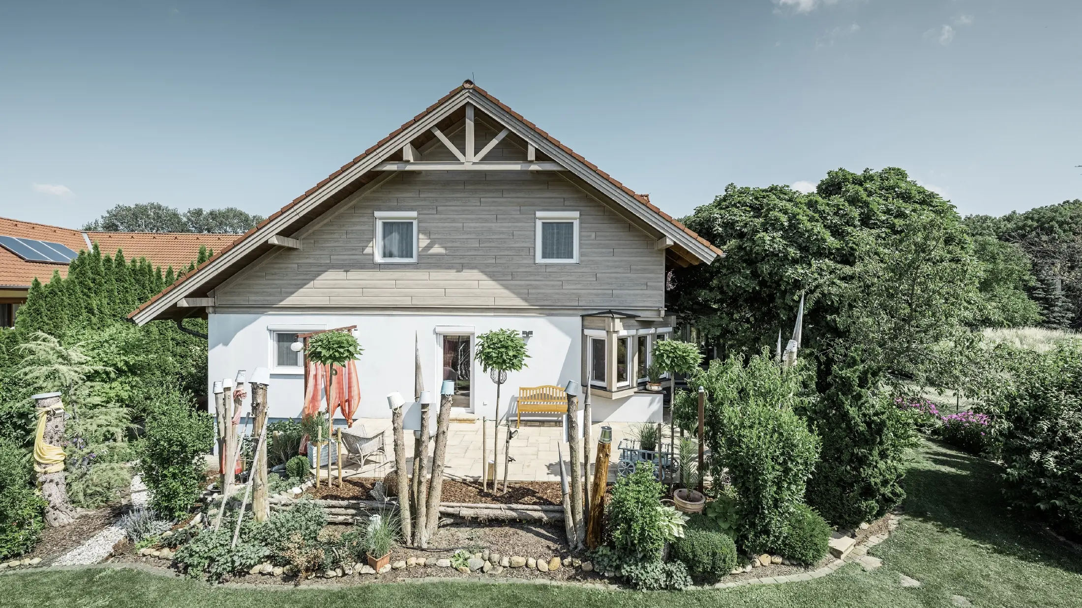 Casa unifamiliare con facciata ristrutturata e rivestimento del frontone rinnovato in un moderno effetto legno nei toni del grigio-beige, abbinata a una facciata intonacata, un tetto a due falde e un giardino ben curato.