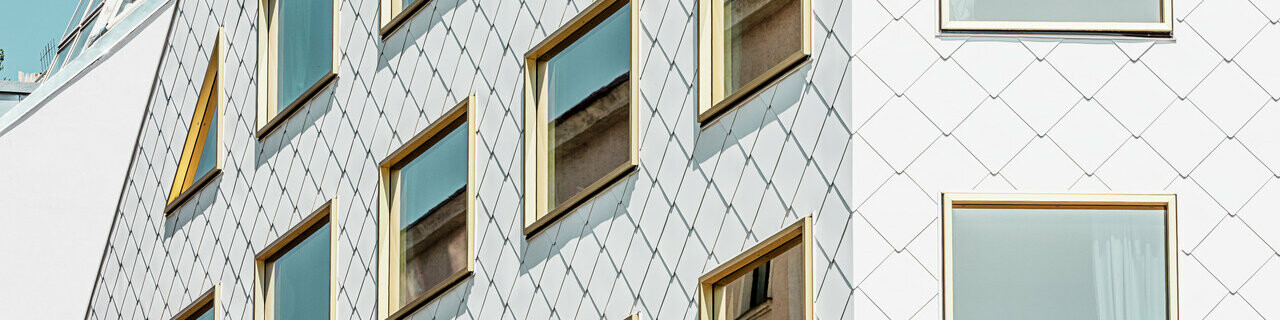 Un moderno hotel a quattro stelle a Vienna, rivestito con i rombi PREFA per tetto e pareti 44 × 44 in P.10 Pure White. La facciata ha una struttura a blocchi con un gran numero di finestre a specchio disposte a griglia, che riflettono i movimenti dell'ambiente circostante.