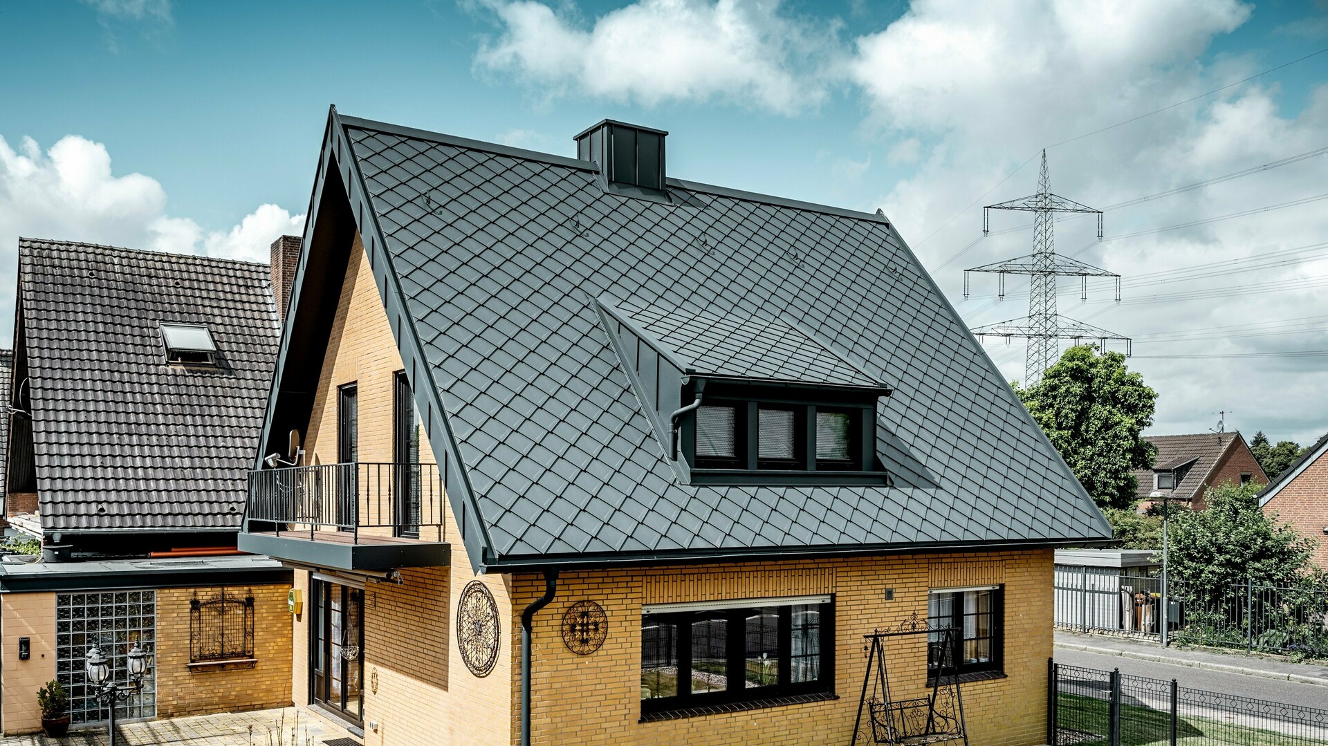 Maison individuelle en briques jaunes après la rénovation du toit avec un losange de toiture PREFA 29 × 29 en gris sombre ; toit en pente raide nouvellement recouvert avec un motif en losange, une lucarne et une cheminée intégrées, terrasse et jardin au premier plan.