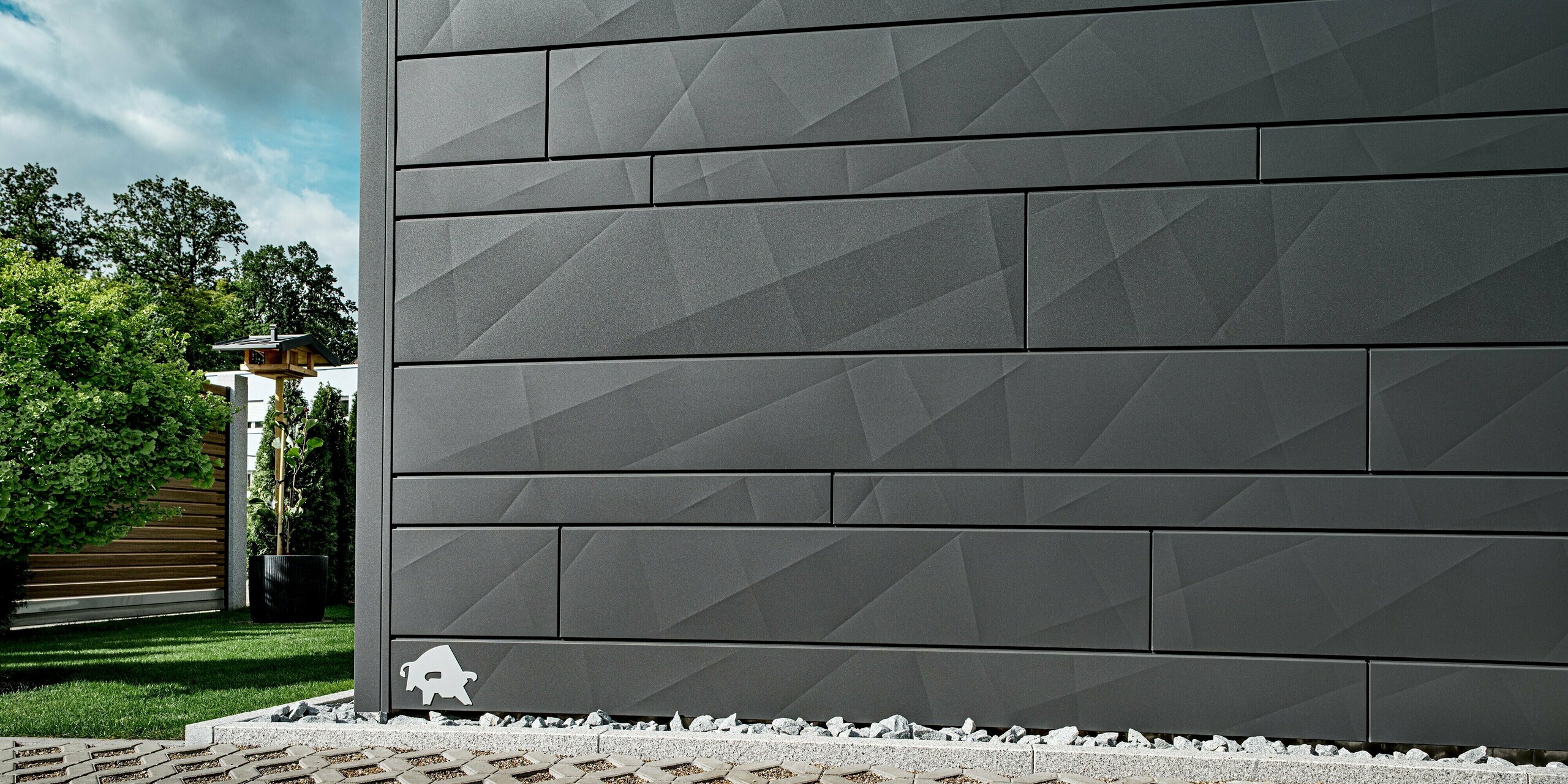 Detailansicht der PREFA Siding.X Aluminiumfassade in der Farbe P.10 Dunkelgrau an einem modernen Einfamilienhaus in Heidenheim. Die klar strukturierten Paneele mit variierenden Breiten und markanten Kantungen erzeugen ein prägnantes, geometrisches Muster, das je nach Lichteinfall spannende Schattenspiele bildet. Im Vordergrund sind helle Ziersteine und ein gepflegter Gartenbereich zu sehen, im Hintergrund Bäume und eine Holzstruktur. Die langlebige und witterungsbeständige Aluminiumoberfläche unterstreicht die hochwertige und moderne Fassadengestaltung. Am linken Rand der Fassade ist das bekannte PREFA-Logo, der Stier, zu erkennen.