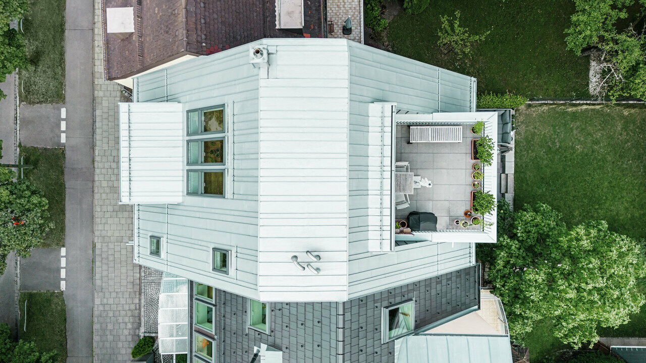 Vue aérienne d'une maison mitoyenne d'angle avec rénovation moderne. La maison dispose d'un toit et d'une façade d'étage en aluminium prépliage métallique argenté, qui souligne les lignes horizontales. Une terrasse sur le toit avec du mobilier d'extérieur et des plantes crée une oasis de verdure urbaine. La façade du sous-sol est d'un ton plus foncé, ce qui crée un contraste attrayant. La maison est entourée d'un espace vert bien entretenu et d'allées pavées qui soulignent la structure claire du bâtiment.
