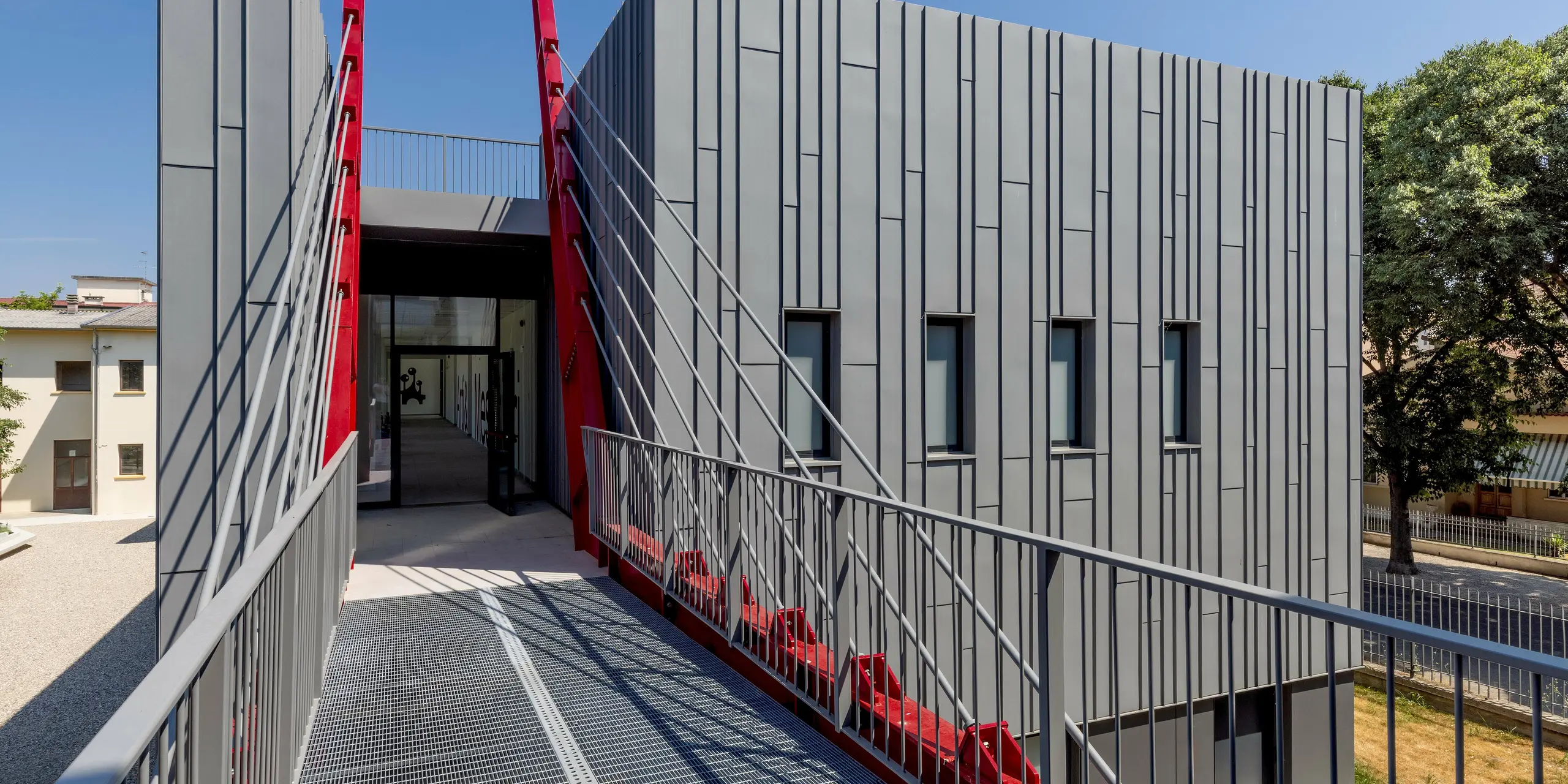 Vue spectaculaire de la Scuola Arte e Mestieri F. Bertazzoni à Suzzara : un pont audacieux, doté d’une grille en acier, mène directement à l’entrée, flanqué de montants diagonaux rouge vif. Ces derniers contrastent avec la façade en aluminium gris sombre, à la structure strictement verticale. La façade PREFALZ confère au bâtiment une surface précise et durable, offrant une résistance maximale aux intempéries. Cet établissement d’enseignement démontre qu’il est possible d’obtenir des résultats exceptionnels en matière de design moderne grâce aux produits en aluminium de haute qualité PREFA.