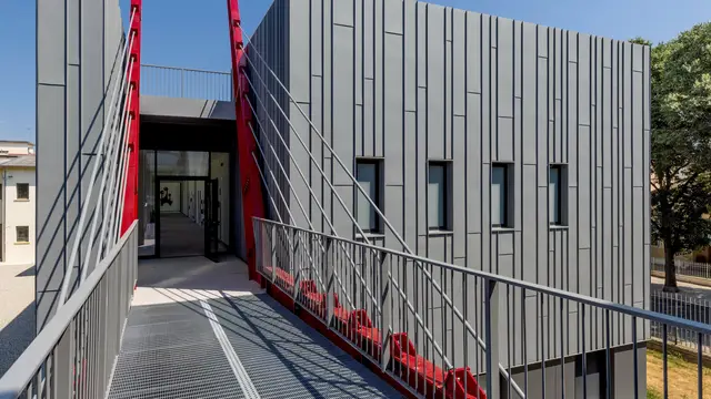 Ein dramatischer Blick auf die Scuola Arte e Mestieri F. Bertazzoni in Suzzara: Eine kühne Brücke mit Stahlgitterrost führt direkt zum Eingang, flankiert von leuchtend roten Diagonalstreben. Diese kontrastieren mit der streng vertikal gegliederten, dunkelgrauen Aluminiumfassade. Die PREFALZ Fassade verleiht dem Bauwerk eine präzise, langlebige Oberfläche mit höchster Witterungsbeständigkeit. Die Bildungseinrichtung beweist, dass sich mit hochwertigen PREFA Aluminiumprodukten spektakuläre Ergebnisse im modernen Design erzielen lassen.