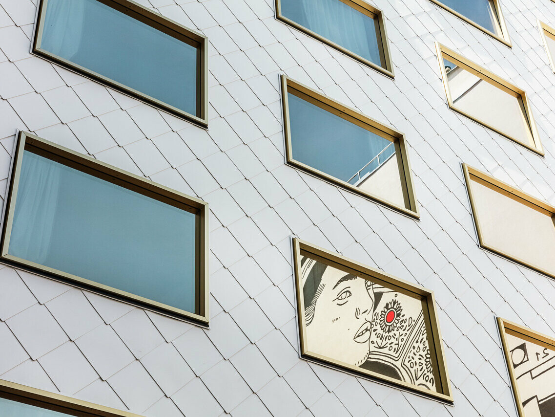 Primo piano della facciata dell'hotel "THE ROCK Radisson RED Vienna", rivestita con tegole PREFA per tetti e pareti 44 × 44 in P.10 Pure White. Tra le numerose vetrate a specchio con cornici dorate, spicca una finestra in cui è incastonata un'opera d'arte che raffigura il ritratto di una persona con un vistoso accento rosso.