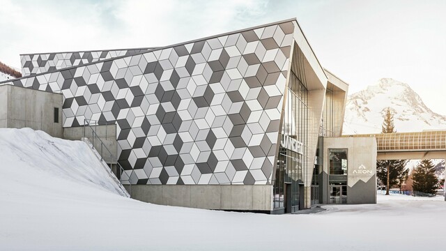 Die Talstation in Les Deux Alpes beeindruckt mit einer außergewöhnlichen Fassade aus PREFABOND Aluminium-Verbundplatten in einem modernen Rautenmuster. Die Kombination aus Weiß- und Grautönen bildet ein markantes Design, das perfekt mit der verschneiten Berglandschaft harmoniert. Die langlebige, wetterbeständige Fassade garantiert auch unter extremen Bergbedingungen eine ästhetische und funktionale Lösung. Die strahlende Sonne im Hintergrund schafft einen majestätischen Rahmen für diese Szenerie.