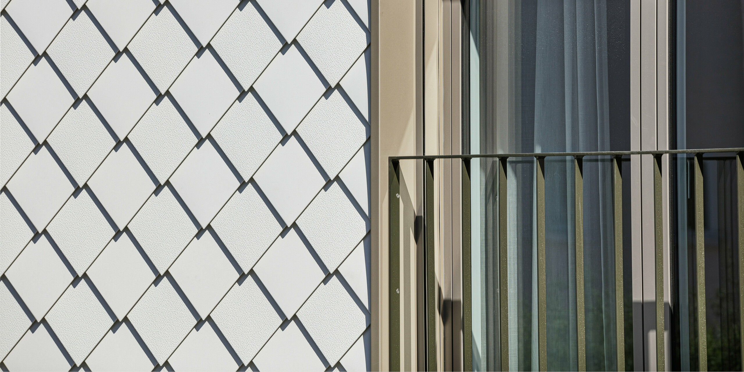 Kontrastreiche Detailaufnahme eines Fensters und der Fassade an einem Wohnhaus in Wiesendangen, Schweiz. Die Fassade zeigt die Verbindung von Funktionalität und Design mit PREFA Wandrauten 20x20 in P.10 Prefaweiß, die neben einem Fenster mit französischem Balkon und eleganten, vertikalen Vorhängen für eine klare und moderne Ästhetik sorgen. Die geometrische Präzision der Rauten schafft eine ansprechende Textur und reflektiert die Qualität und Innovationskraft der PREFA Aluminiumprodukte.