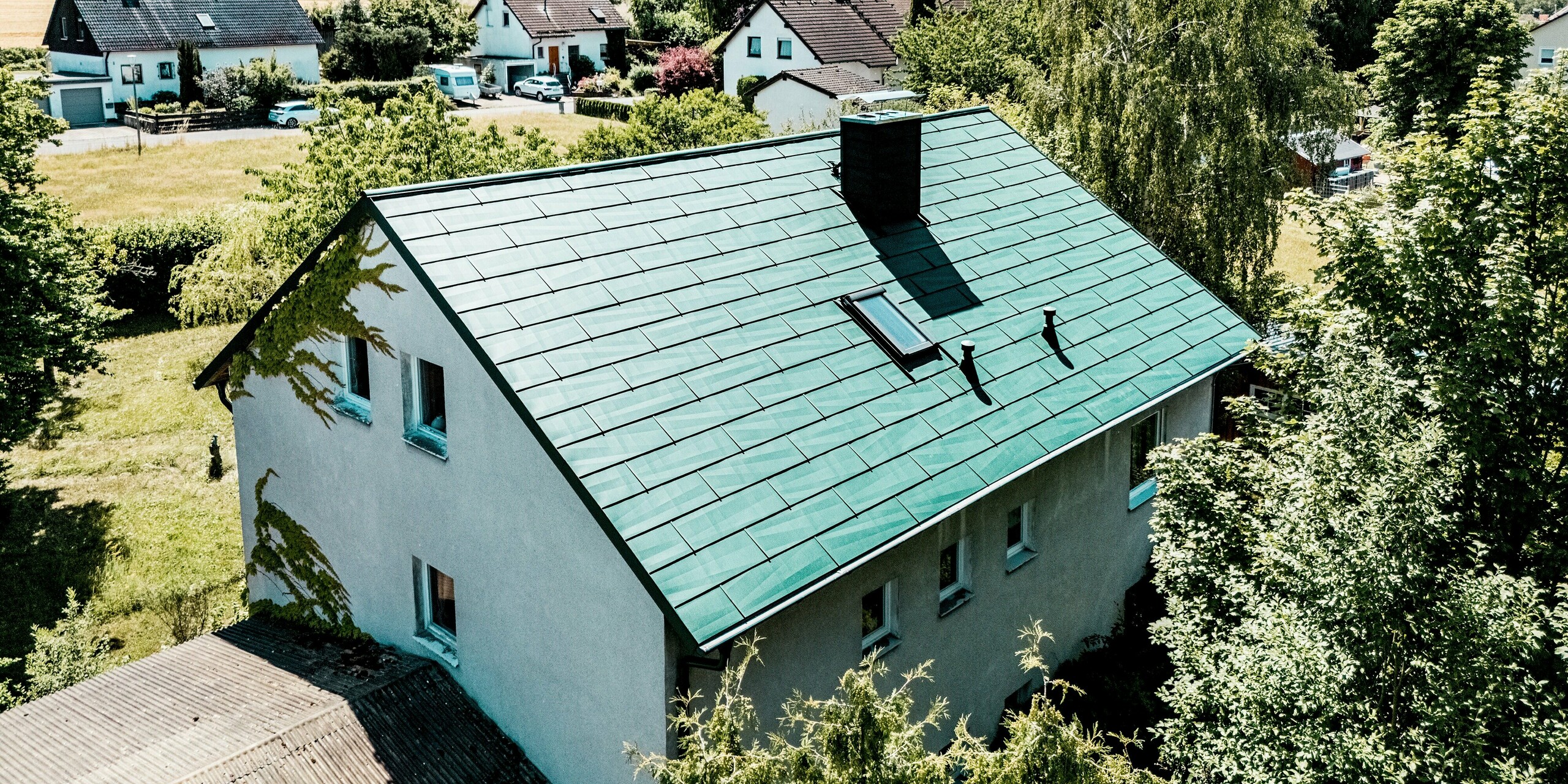 Maison individuelle à Schwarzenbach-Martinlamitz avec une toiture modernisée en panneaux PREFA FX.12 couleur Vert Mousse P.10. La photo met en valeur la toiture rénovée, avec ses lignes épurées, son puits de lumière remarquable et sa cheminée élégamment intégrée, le tout sur fond de paysage champêtre. La toiture en aluminium séduit par son aspect haut de gamme et les bords irréguliers des panneaux, qui reflètent magnifiquement la lumière. Résistante aux intempéries, durable et sans entretien, la toiture PREFA offre une protection fiable et confère à la maison une allure moderne et distinctive au cœur de la verdure.