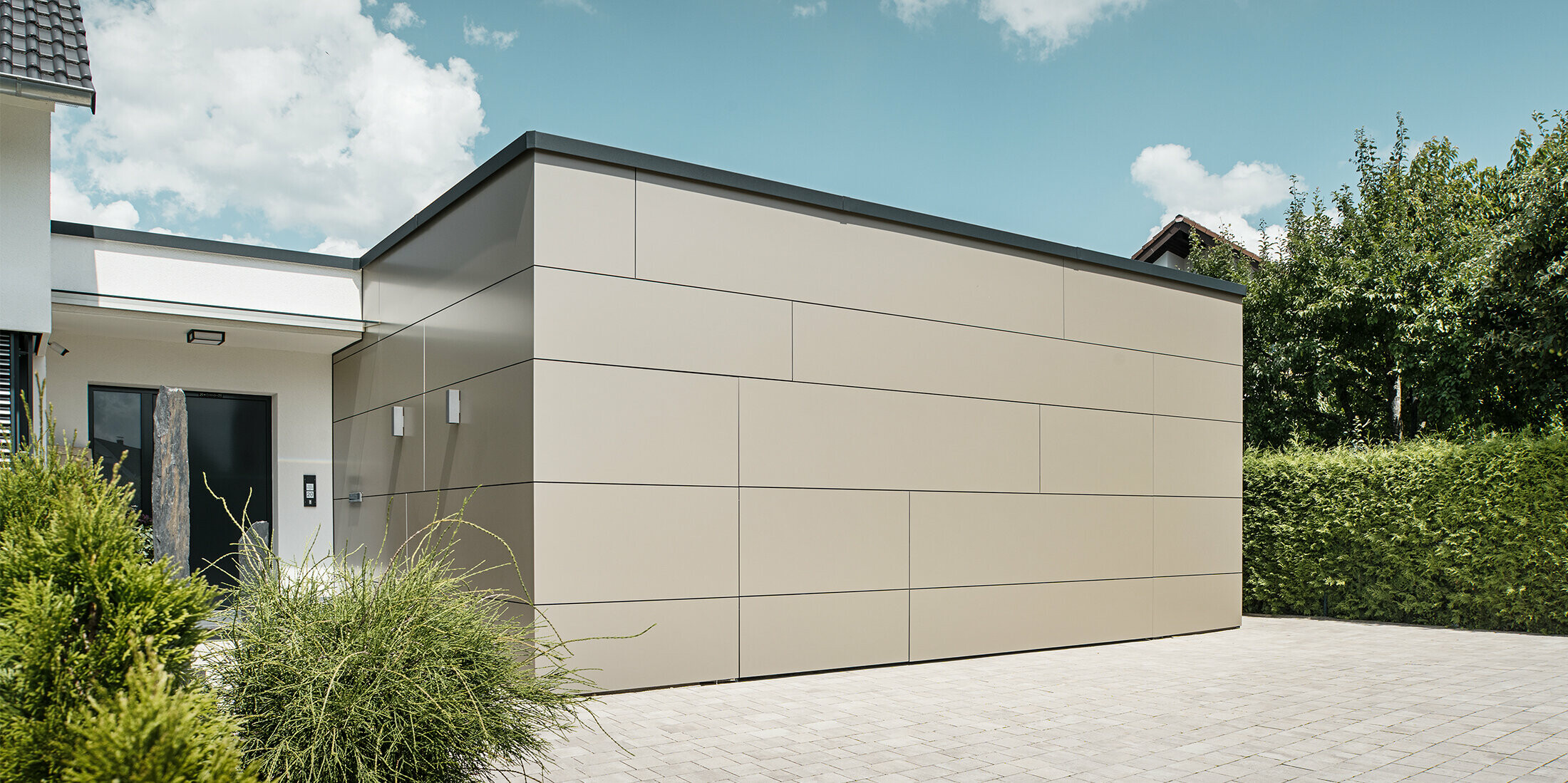 Gesamtansicht einer Garage im geschlossenen Zustand. Das geschlossene Garagentor lässt die Öffnungsmöglichkeit nur schwer erkennen, da die PREFABOND Aluminium Verbundplatten in Bronze das Gebäude fast nahtlos verkleiden. Die Pflanzen vor der Eingangstür zeugen von der Pflege des schönen Einfamilienhauses.