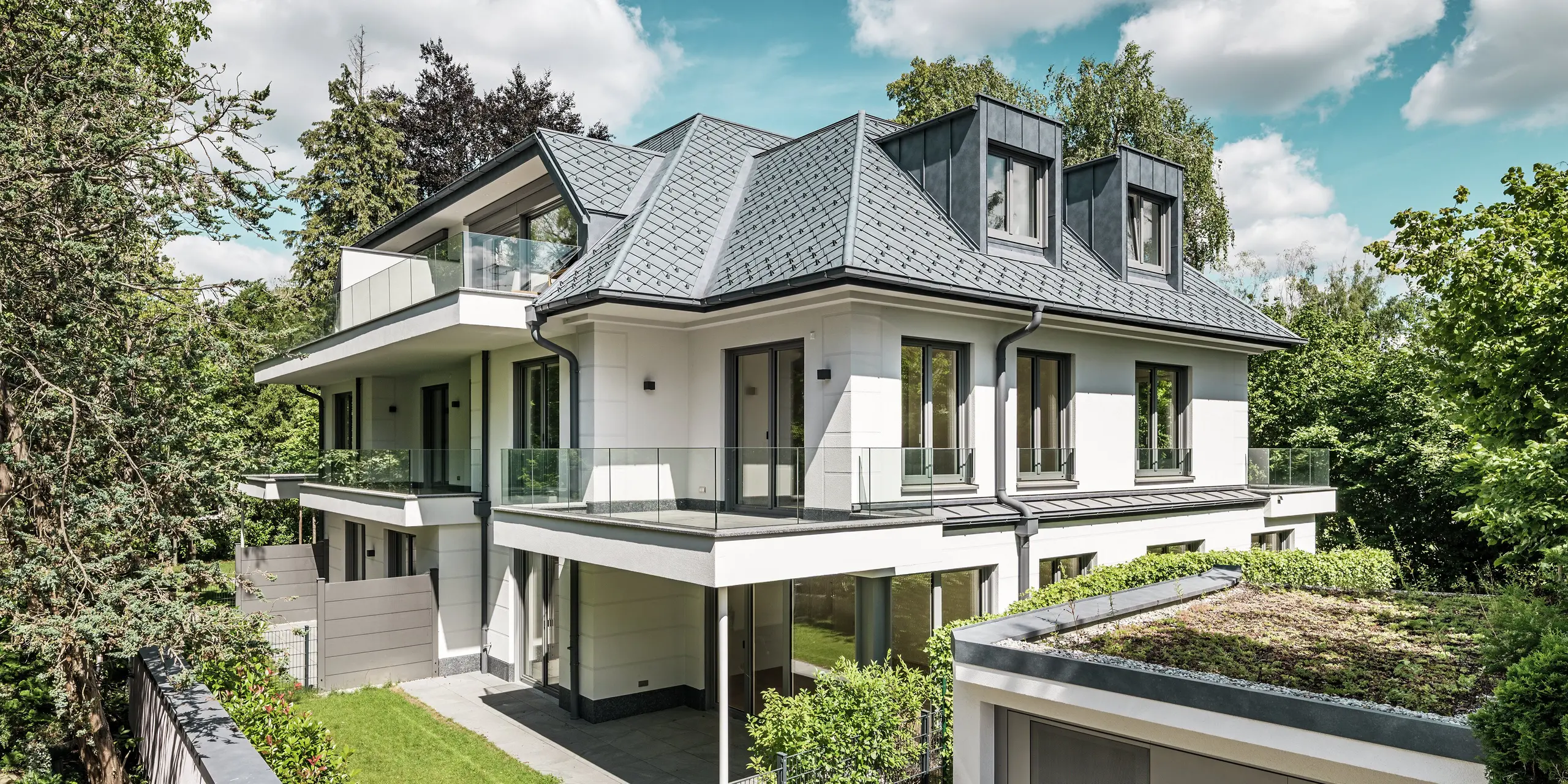 Vue d'un immeuble d'appartements moderne au design élégant près de Munich. La toiture est recouverte de losanges PREFA 29 x 29 cm, coloris Gris Pierre P.10. La charpente remarquable est mise en valeur par d'élégantes lucarnes plates en PREFALZ, à la fois esthétiques et fonctionnelles. Le jardin soigné et la vaste terrasse soulignent la qualité architecturale et créent une harmonie entre intérieur et extérieur. Résistants et durables, les produits PREFA s'intègrent parfaitement au paysage verdoyant et offrent une protection et une beauté pérennes.