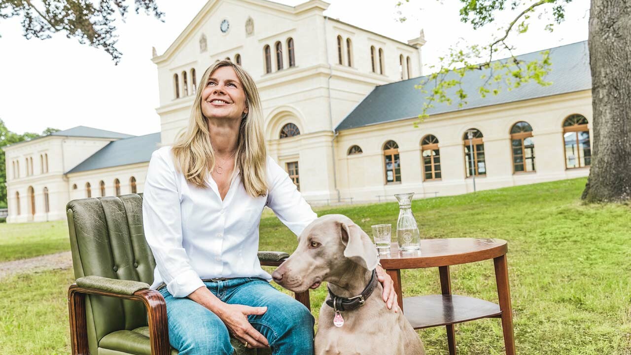 Aufnahme von Sabine Rothe beim Interview zur Sanierung des Marstalls in Basedow, Mecklenburg-Vorpommern, mit ihrem Hund vor dem Schloss mit den neu verlegten Dachschindeln von PREFA in Anthrazit.