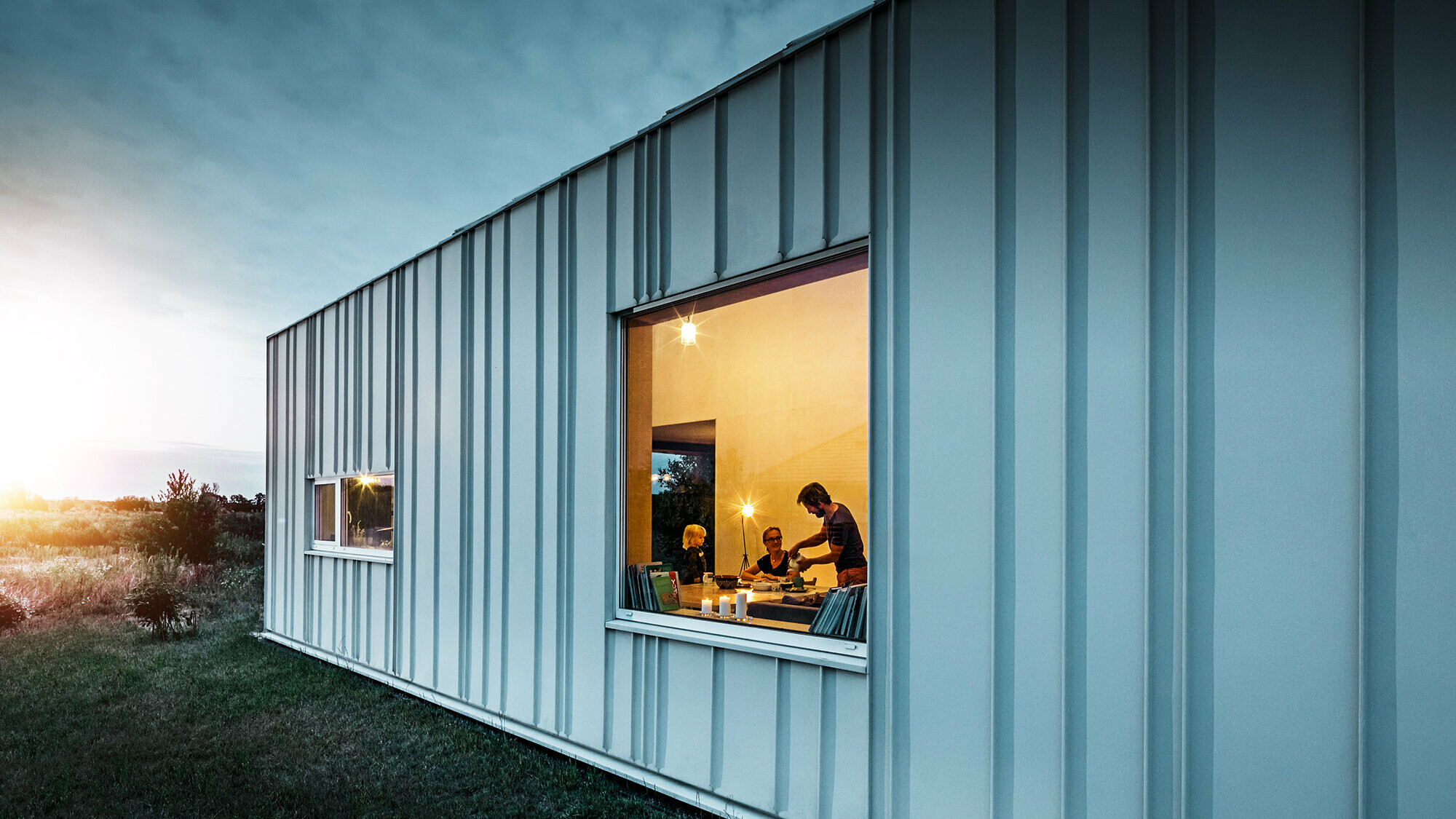 Winkelstehfalz Fassade mit PREFALZ in unterschiedlichen Scharenbreiten in P.10 Prefaweiß bei Dämmerung fotografiert. Durch das Esszimmer erkennt man eine Familie beim Abendessen. 