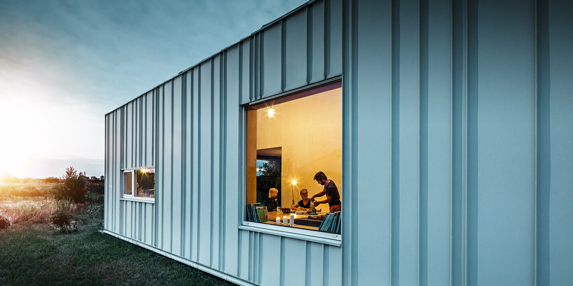 Façade avec joint debout et différentes largeurs de lames en PREFALZ de P.10 blanc PREFA photographiée au crépuscule. À travers la fenêtre de la salle à manger, on peut voir une famille en train de dîner.