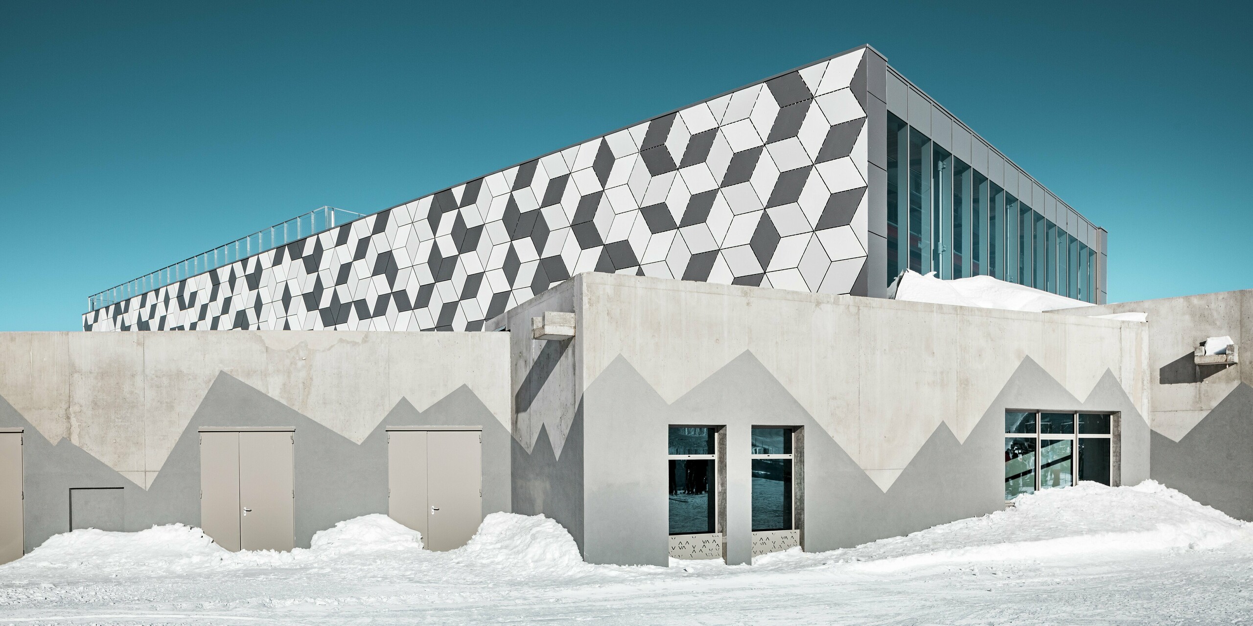 Die moderne Bergstation der Seilbahn 3S in Les Deux Alpes glänzt mit einer futuristischen Fassade aus PREFABOND Aluminium Verbundplatten in auffälliger Rautenoptik. Die Farben P.10 Prefaweiß, P.10 Reinweiß und Rauchsilber erzeugen ein faszinierendes 3D-Muster, das sich perfekt in die verschneite Hochgebirgslandschaft einfügt. Die Fassade ist witterungsbeständig, langlebig und ästhetisch ein echtes Highlight. Der untere Teil des Gebäudes besteht aus Sichtbeton. Mit dunkelgrauer Farbe wurden hier Berge nachgemalt.