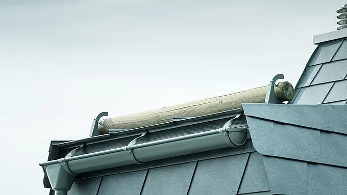 Vue du toit d'un chalet en bois au bord du lac Léman avec des bardeaux PREFA gris pierre, une gouttière moderne et une lucarne. L'image montre une combinaison parfaite entre design fonctionnel et élégance esthétique, idéale pour une architecture durable.
