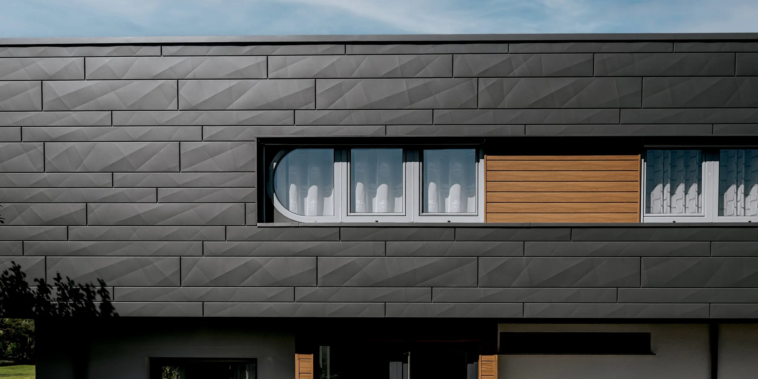 Detailansicht der modernen PREFA Aluminiumfassade am Einfamilienhaus in Marksuhl: Die markante Oberfläche der PREFA Siding.X Paneele in P.10 Dunkelgrau erzeugt ein faszinierendes Spiel aus Licht und Schatten. Warme Siding-Elemente in Walnuss braun setzen natürliche Akzente. Die robuste Aluminiumverkleidung überzeugt mit langlebigem Schutz, Witterungsbeständigkeit und zeitloser Ästhetik.
