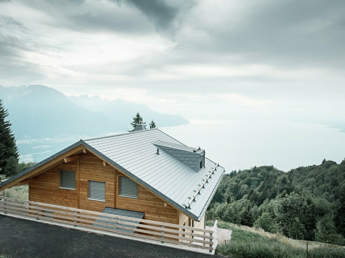 Moderne Holz-Lodge in der Schweiz am Genfersee mit PREFA Dachschindeln in der Farbe Steingrau, umgeben von grünen Wäldern und Berglandschaften. Perfekte Kombination aus natürlichem Holz und langlebigem Aluminiumdach für nachhaltigen Wohnkomfort.