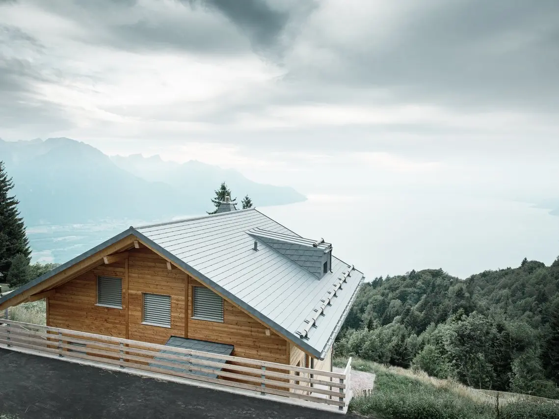 Lodge moderne en bois en Suisse, au bord du lac Léman, avec des bardeaux PREFA de couleur gris pierre, entourée de forêts verdoyantes et de paysages montagneux. Combinaison parfaite entre le bois naturel et un toit en aluminium durable pour un confort de vie durable.