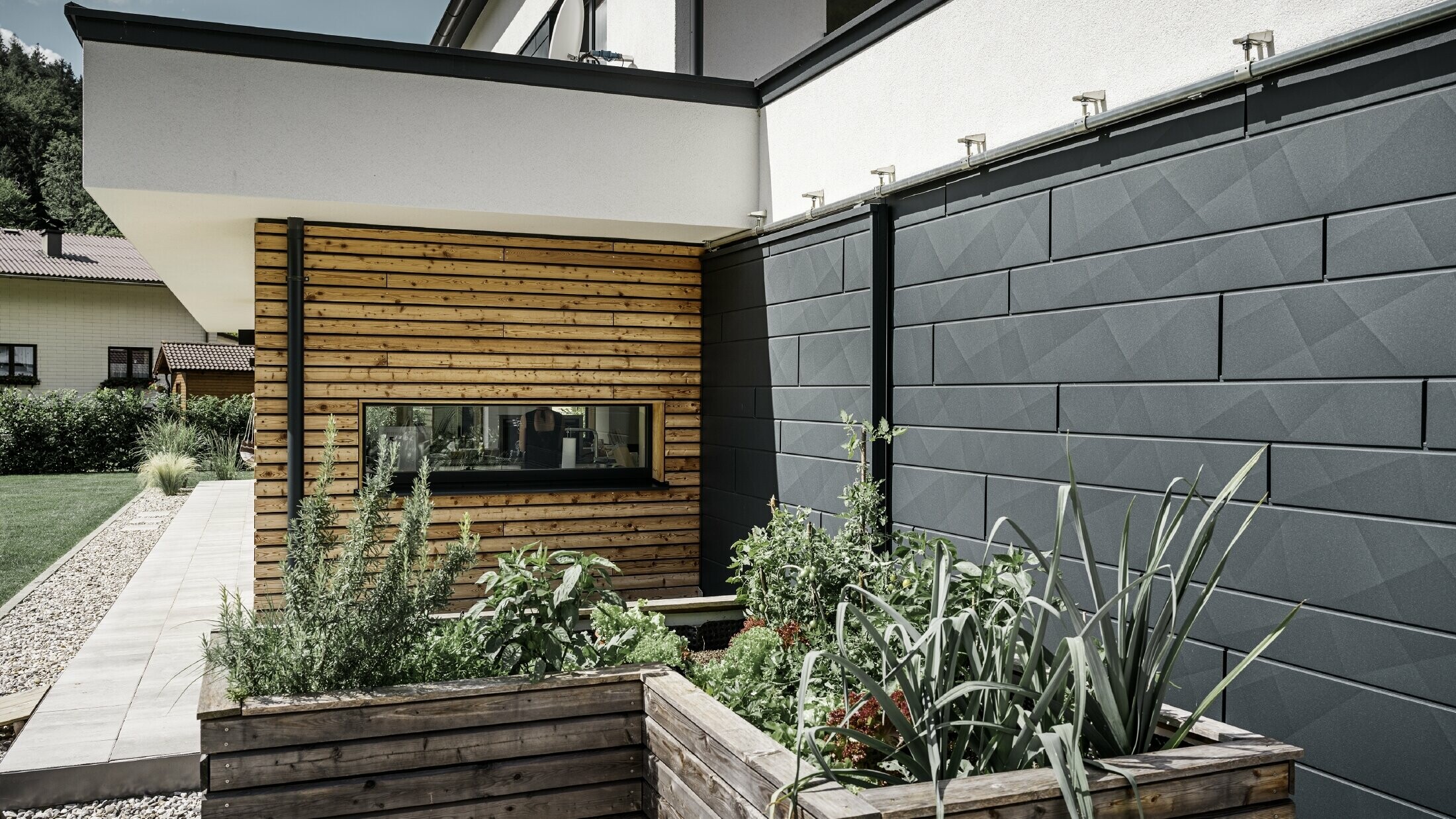 Dieses Bild zeigt eine Nahaufnahme des Einfamilienhauses, bei dem die Fassadenverkleidung mit PREFA Siding.X in P.10 Anthrazit im Fokus steht. Die dunklen Fassadenpaneele mit der markanten Knitteroptik bieten einen ansprechenden visuellen Kontrast zu den lebendigen, holzverkleideten Bereichen und den umgebenden Grünflächen. Vor der Fassade aus Aluminium sind Hochbeete positioniert, die mit verschiedenen Pflanzen und Sträuchern bepflanzt sind, was die natürliche und nachhaltige Ausstrahlung des Hauses unterstreicht. Die Kombination aus modernen Materialien und der Integration in den Gartenbereich vermittelt ein Bild von zeitgemäßer Architektur, die sich harmonisch in ihre Umgebung einfügt.