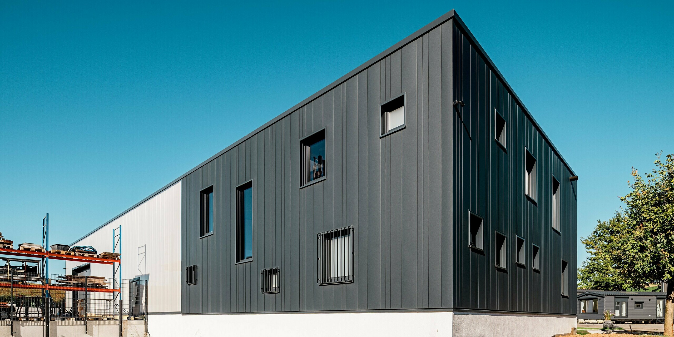 Dieses Bild zeigt die Rückseite des Firmengebäudes der Gebäudetechnik Schädel GmbH, wo die vertikale Linienführung der PREFA Sidings besonders deutlich zur Geltung kommt. Die kräftige P.10 Anthrazit Oberfläche verleiht dem kompakten Baukörper Stabilität und Eleganz. Dank der verschiedenen Paneelbreiten entsteht ein lebendiges Fassadenbild. Die PREFA Aluminiumfassade schützt nachhaltig vor Witterungseinflüssen und bleibt wartungsarm.