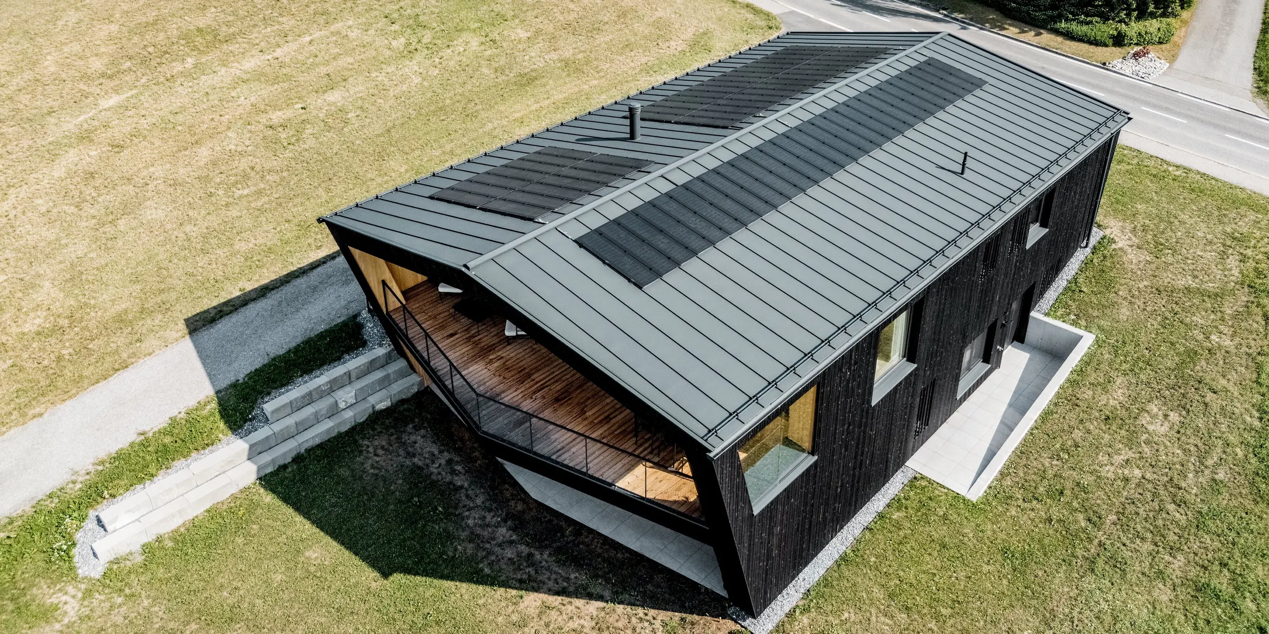 Spektakuläre Vogelperspektive auf das Einfamilienhaus in Röthis: Das markante Satteldach mit PREFALZ in P.10 Anthrazit und harmonisch integrierten Solarmodulen PREFALZ prägt die moderne Architektur. Die dunkle Holzfassade und der geschützte Balkon setzen spannende Akzente. Die leichte, langlebige und witterungsbeständige Aluminiumdeckung verbindet elegantes Design mit nachhaltiger Energiegewinnung.