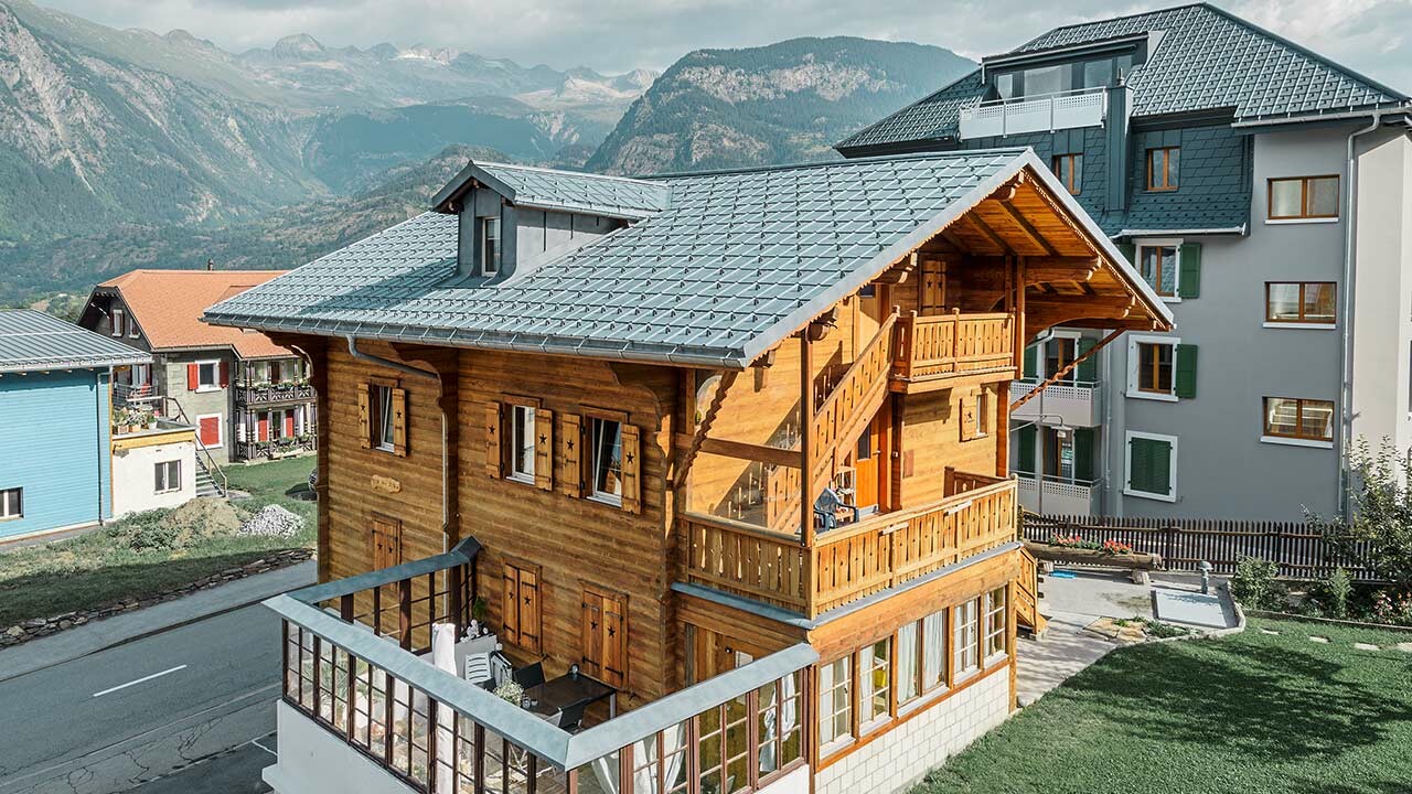 Chalet en bois suisse traditionnel avec lucarne et toit à deux pans ; la toiture a été recouverte de tuiles PREFA classiques couleur gris pierre.