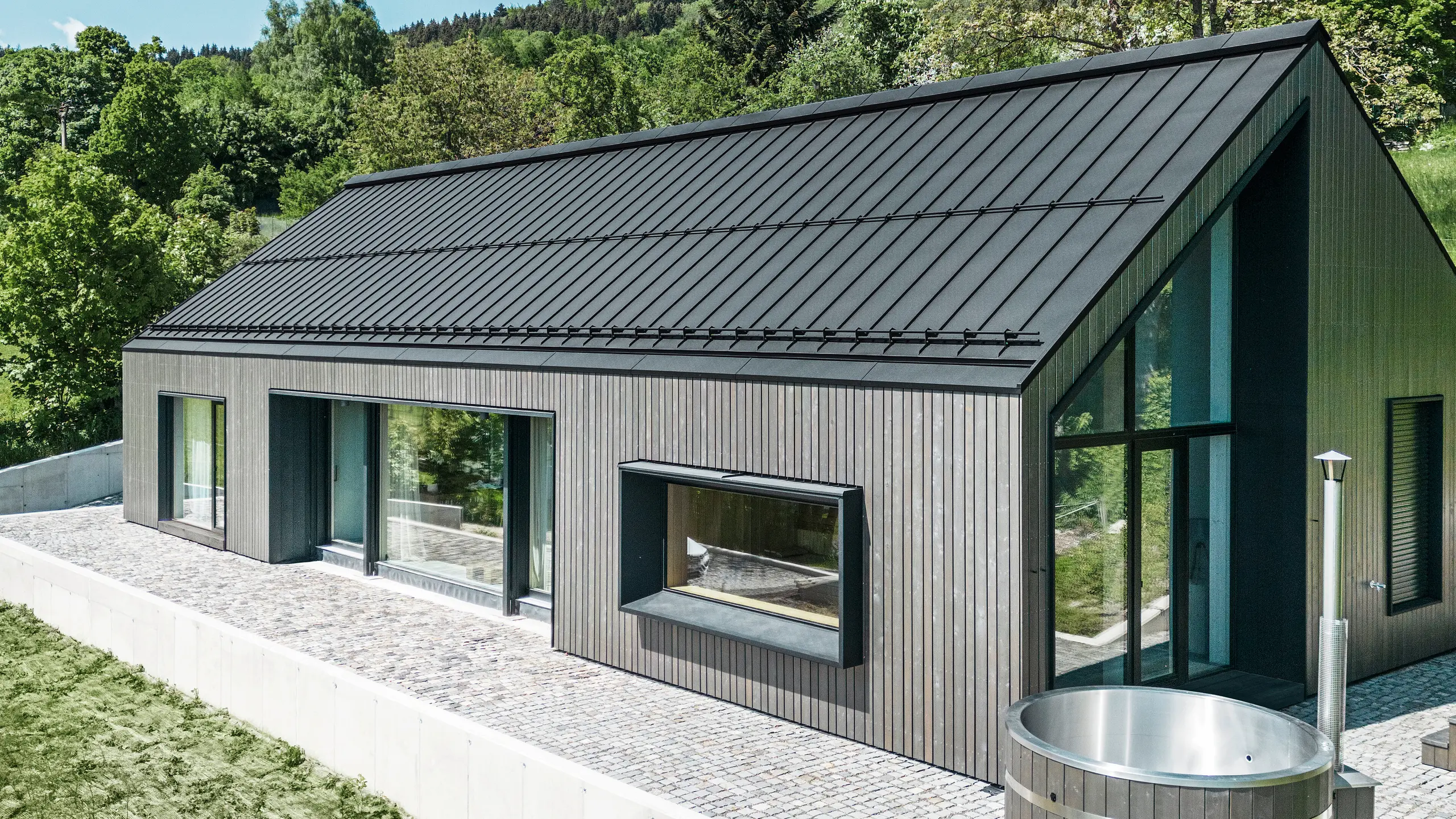 Ein modernes Holzmassivhaus in Kuncice (CZ) wurde mit großen Fensterflächen ausgestattet. Das dunkle Holzmassivhaus verfügt über ein Dachsystem aus PREFALZ in der Farbe P. 10 Schwarz. Im hinteren Außenbereich steht ein Whirlpool mit einer Holzverkleidung.
