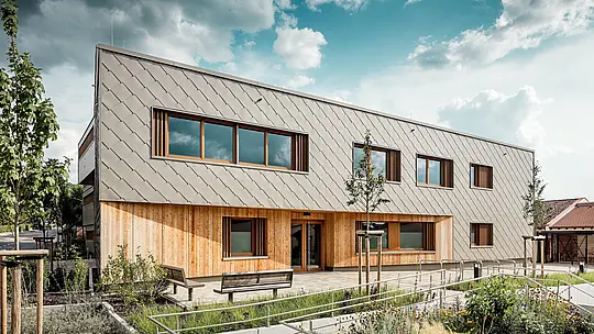 Die frontal aufgenommene Ansicht der Kindertagesstätte in Alzenau zeigt den einladenden Haupteingang mit seiner harmonischen Kombination aus bronzefarbenen PREFA Wandrauten 44 × 44 und vertikaler Holzfassade im Erdgeschoss. Die klar strukturierte Aluminiumverkleidung erzeugt ein elegantes Schuppenmuster, das durch großzügige Fenster mit Holzrahmen und vertikalen Lamellen rhythmisch unterbrochen wird. Im Vordergrund unterstreichen barrierefreie Wegeführung, Blumenbeete und Sitzbänke die hohe Aufenthaltsqualität. Das Zusammenspiel aus langlebigem Aluminium, natürlichem Holz und durchdachter Freiraumgestaltung macht das Gebäude zu einem architektonischen Vorzeigeprojekt für nachhaltige, funktionale und ästhetisch hochwertige Bildungsbauten mit PREFA Fassadensystemen.