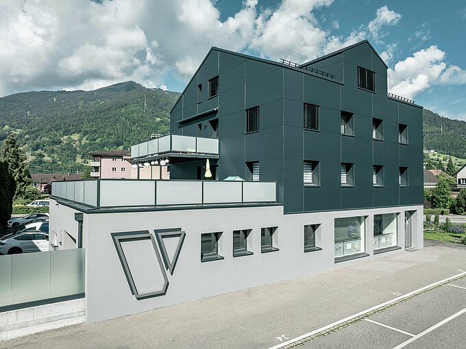 Das Architekturbüro Voser in Landquart, im Schweizer Graubünden, mit einer PREFA Aluminiumfassade in Anthrazit. Das Gebäude wurde saniert und mit PREFABOND Aluminium Verbundplatten ausgestattet. Die Gestaltung kombiniert klare Linien und hochwertige Materialien. Auffällig sind die großen Balkone mit Glasbrüstungen und das prägnante Logo des Architekturbüros Voser an der Gebäudeseite. Im Hintergrund ist eine grüne Berglandschaft zu erkennen.