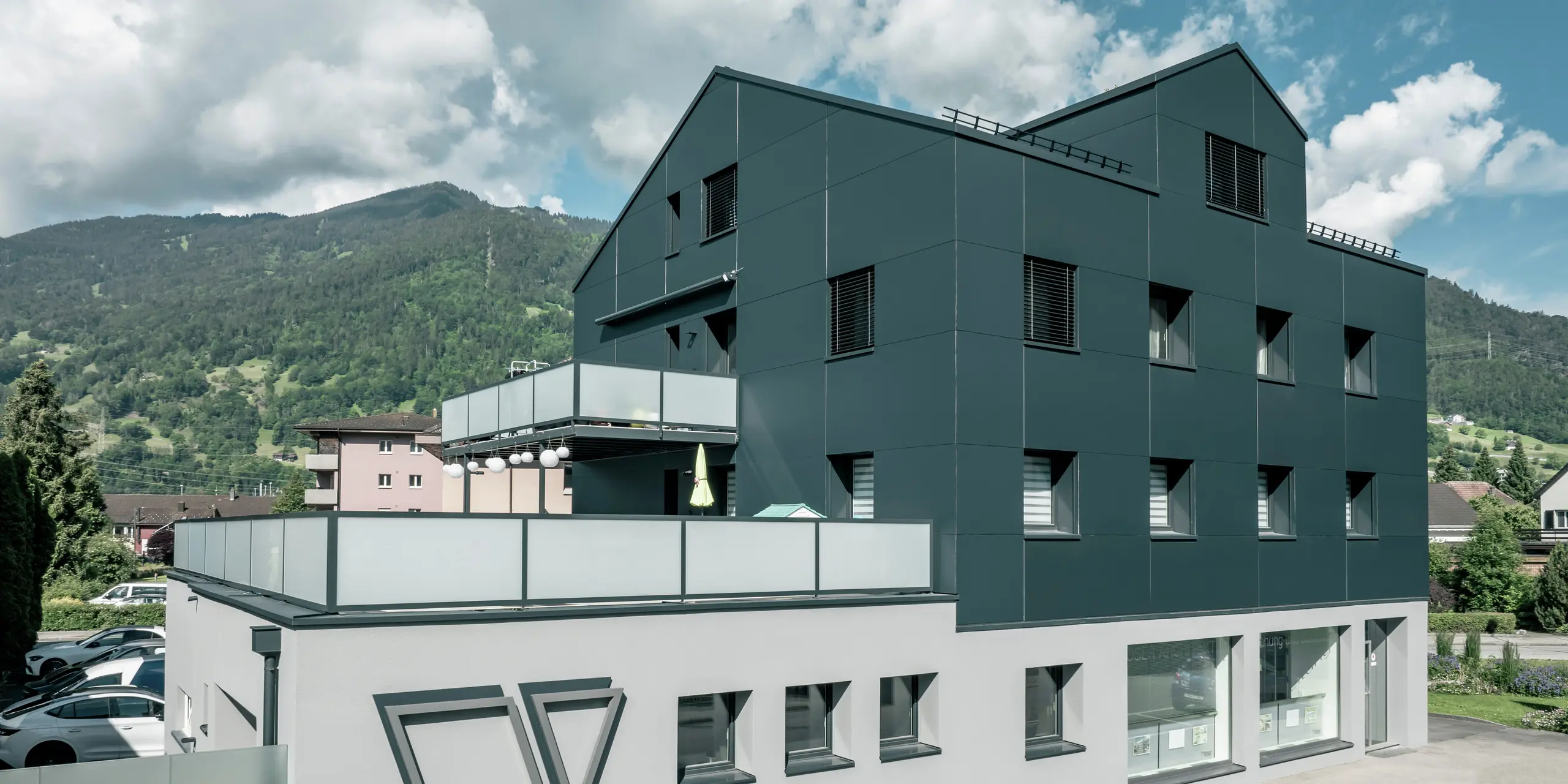 Le bureau d'architecture Voser à Landquart, dans le canton suisse des Grisons, avec une façade en aluminium PREFA en anthracite. Le bâtiment a été rénové et équipé de panneaux composites en aluminium PREFABOND. Le design allie des lignes claires et des matériaux de haute qualité. Les grands balcons avec parapets en verre et le logo frappant du cabinet d'architectes Voser sur le côté du bâtiment sont frappants. Un paysage de montagne verdoyant peut être vu en arrière-plan.