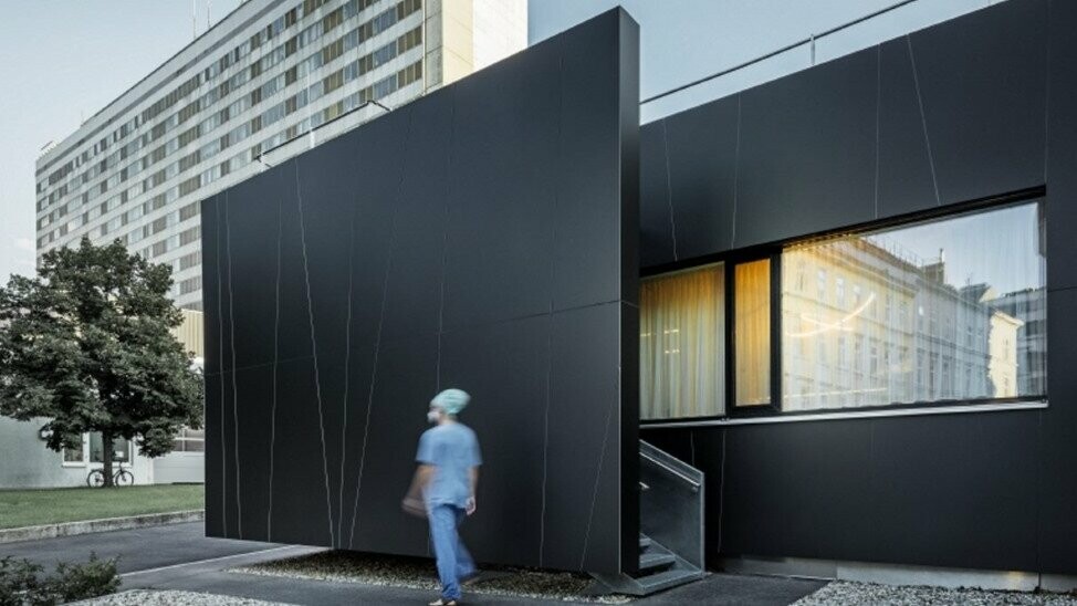 Frontalansicht der zentralen Notaufnahme der Klinik Landstraße in der Juchsgasse in Wien wurde mit PREFABOND Aluminium Verbundplatten in der Farbe schwarzgrau verkleidet. Im Bild ist eine Krankenschwester zu sehen.