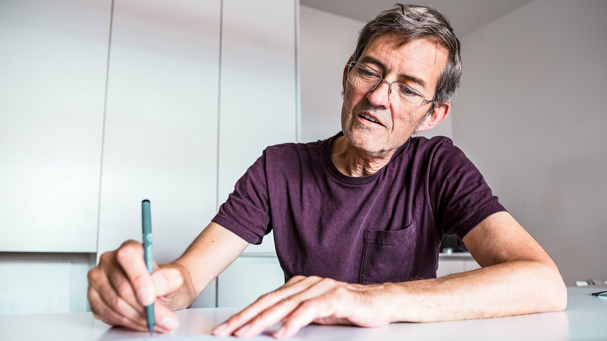 L'architecte Dominique Cornaert est assis à une table blanche et dessine. Derrière lui, on peut voir des murs blancs ainsi qu'une armoire blanche.