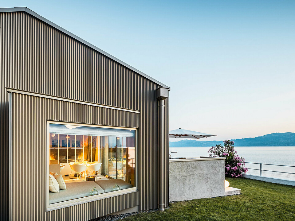 Vista laterale della piccola casa dell'architetta Sophie Morard caratterizzata da una facciata in alluminio con profilo a zeta direttamente sul lago di Ginevra. Si scorge una parte degli interni illuminati.