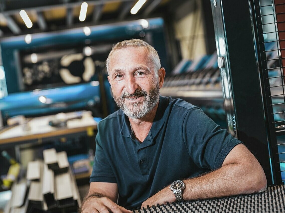 Roger Emmenegger, maestro lattoniere presso Pilatus Flachdach AG, sorride alla telecamera. È seduto in un moderno laboratorio con macchinari e materiale sullo sfondo. Emmenegger indossa una polo blu scuro e un orologio da polso, che sottolineano l'atmosfera rilassata e professionale.